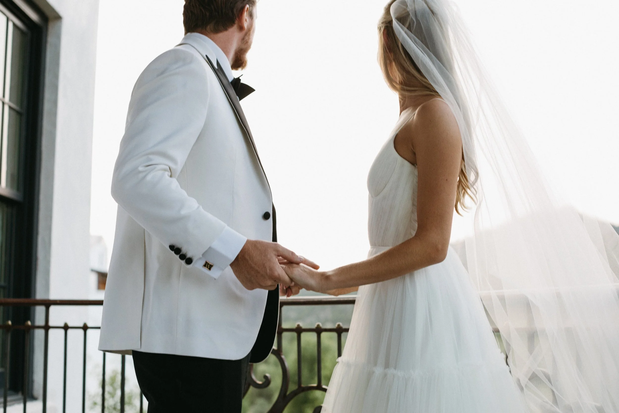 A bride and groom holding hands during a wedding ceremony on a balcony with a scenic background.