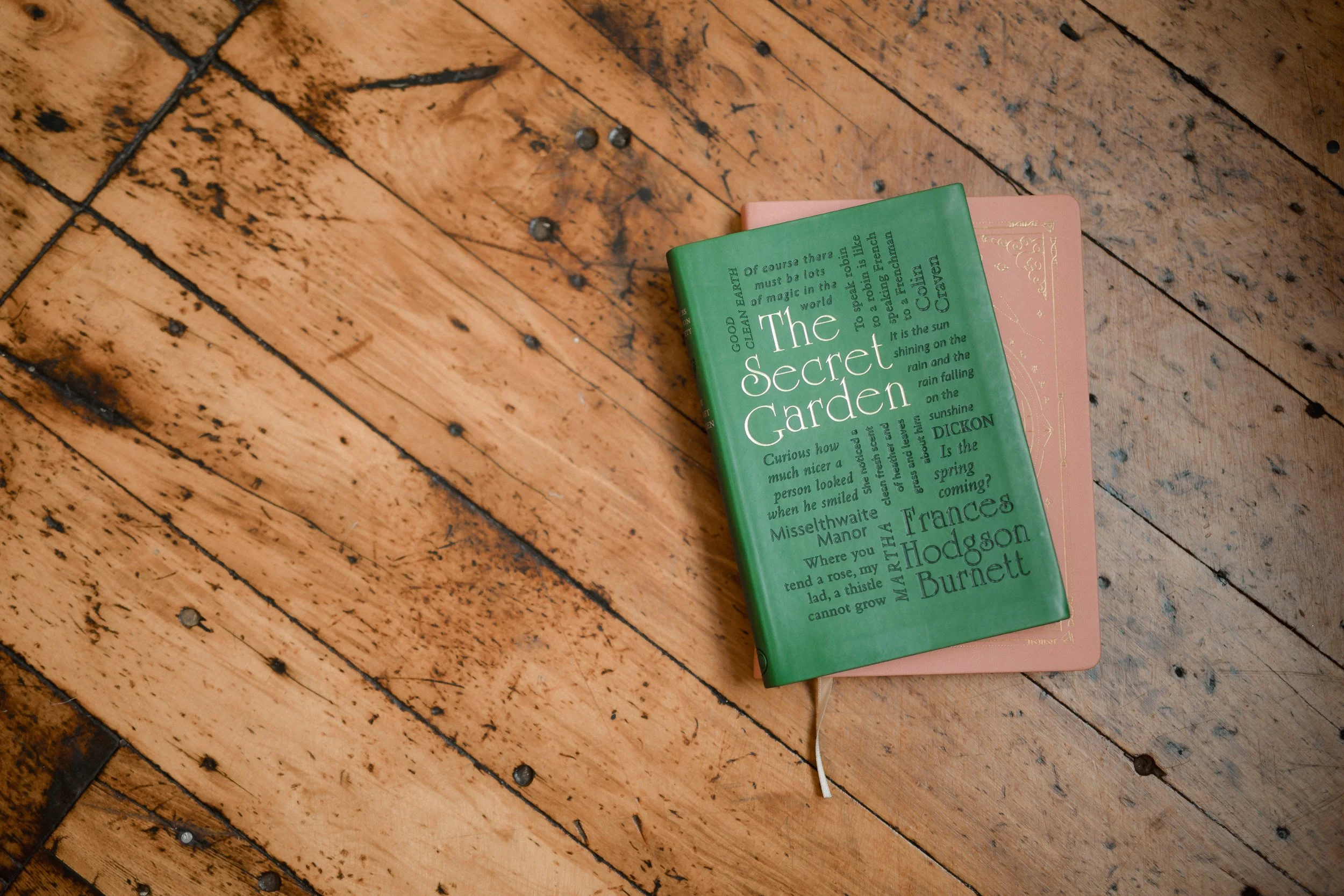 A green hardcover book titled 'The Secret Garden' lying on top of a pink hardcover book, both placed on a wooden floor.