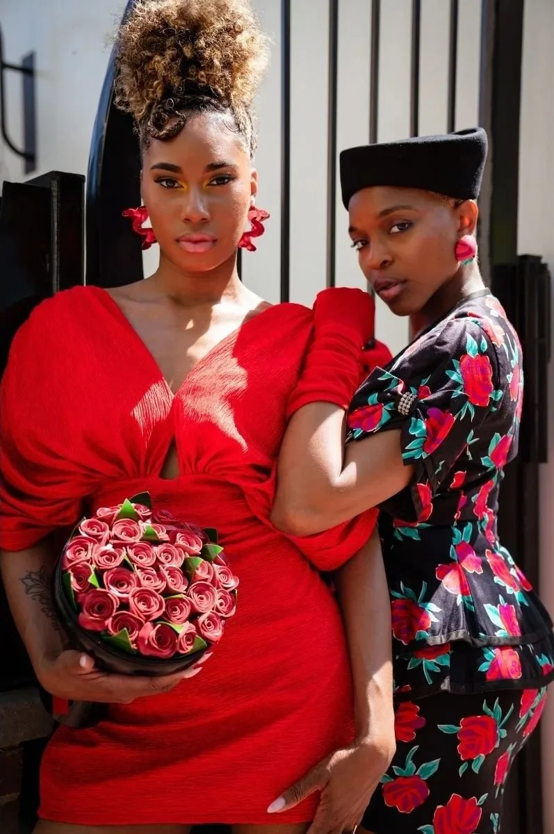 Two women standing close together, one holding a bouquet of pink roses. The woman on the left is wearing a bright red dress with puffed sleeves, curly hair styled in an updo, and bold earrings, while the woman on the right is dressed in a black floral dress with a matching hat and earrings, standing in front of a black and white striped background.