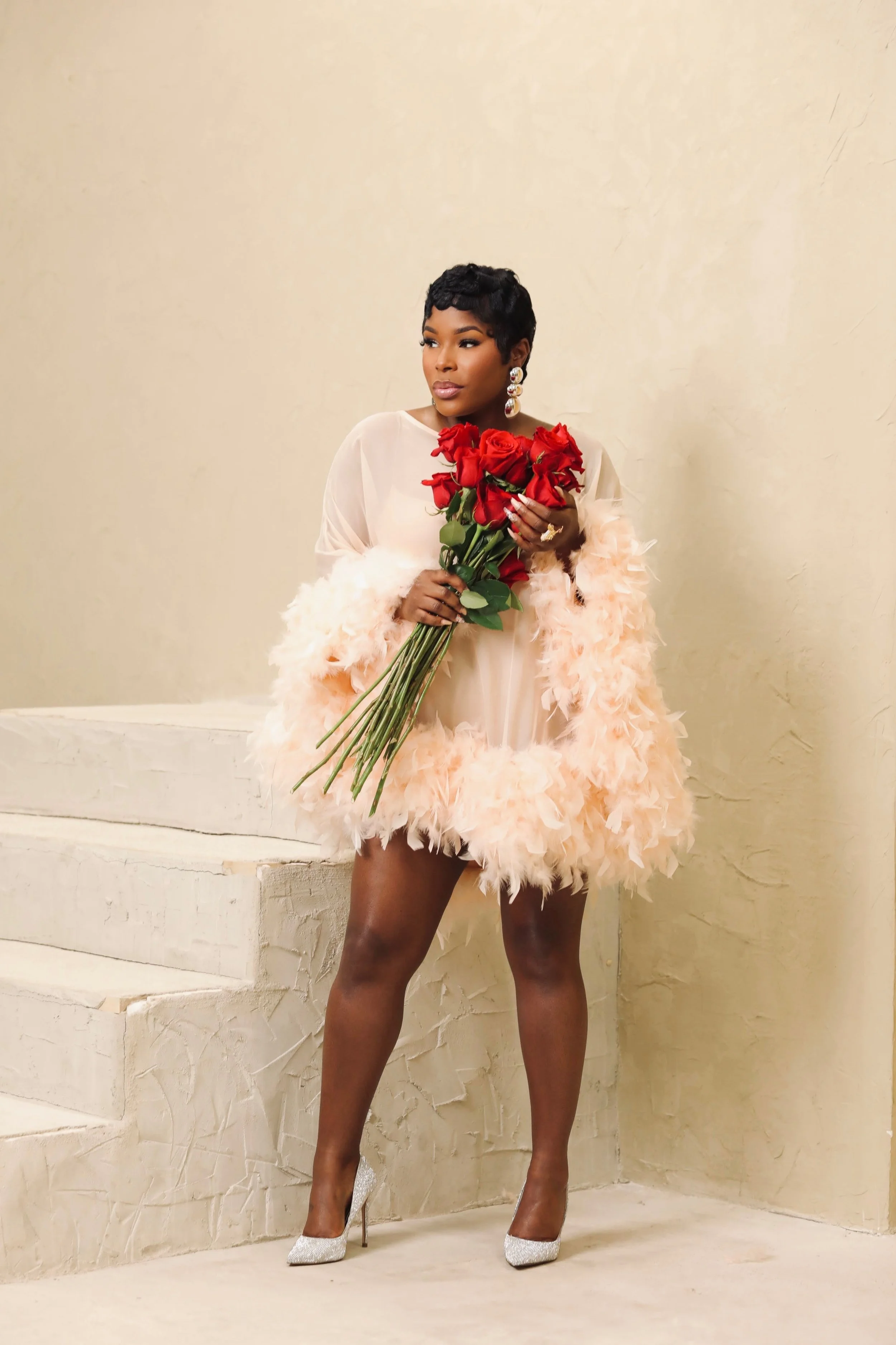 A woman with short black hair holding a bouquet of red roses, wearing a light-colored dress with feathered embellishments and sparkling high heels, standing against a textured beige wall.