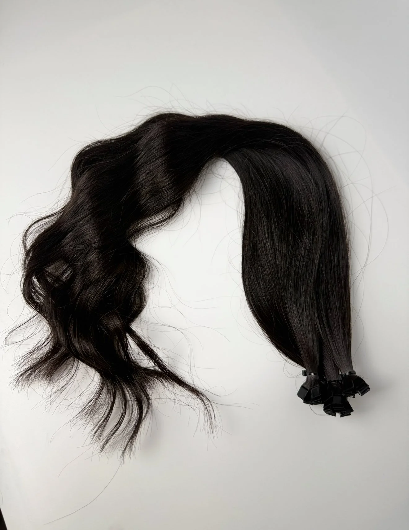 Black hair extensions, wavy on the left side and straight on the right side, tied with black clips at the ends, laid on a white surface.
