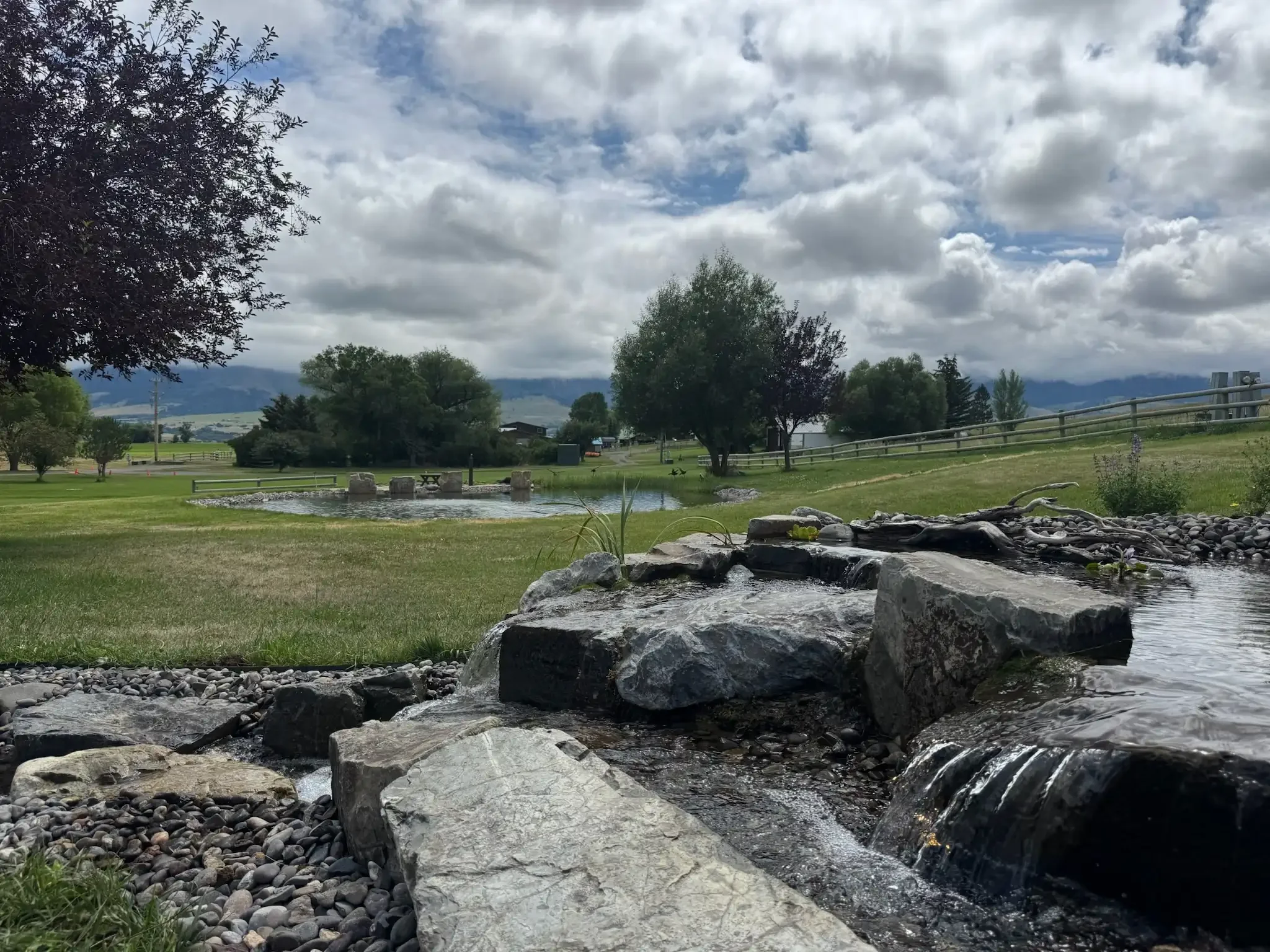 A water stream flowing over boulders into a waterfall with a glistening pond in the back ground.