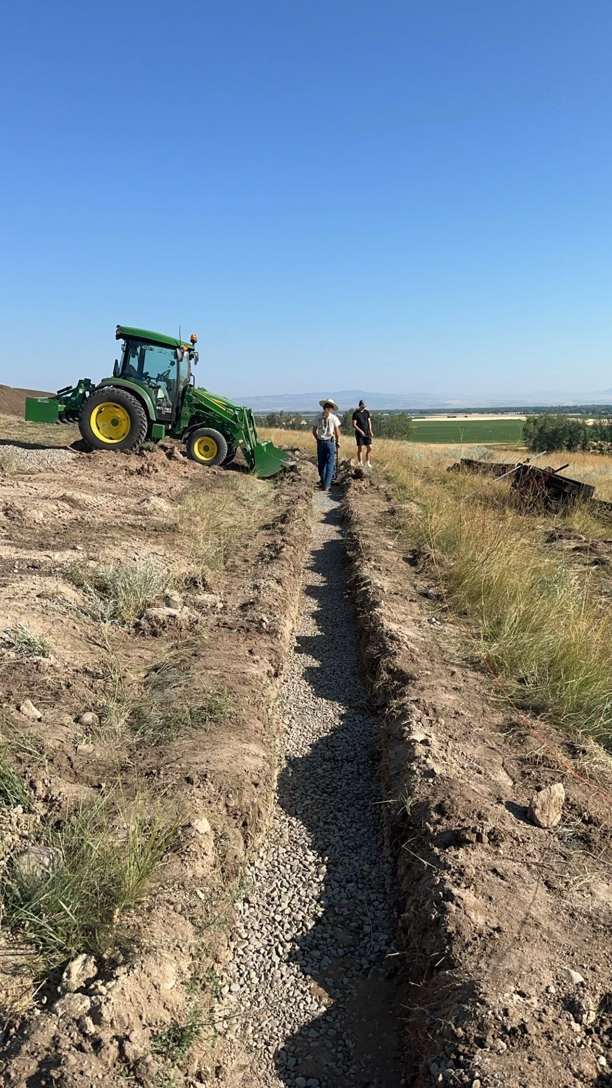 Two men working on a farm field with a green tractor and a long trench filled with gravel, under a clear blue sky.