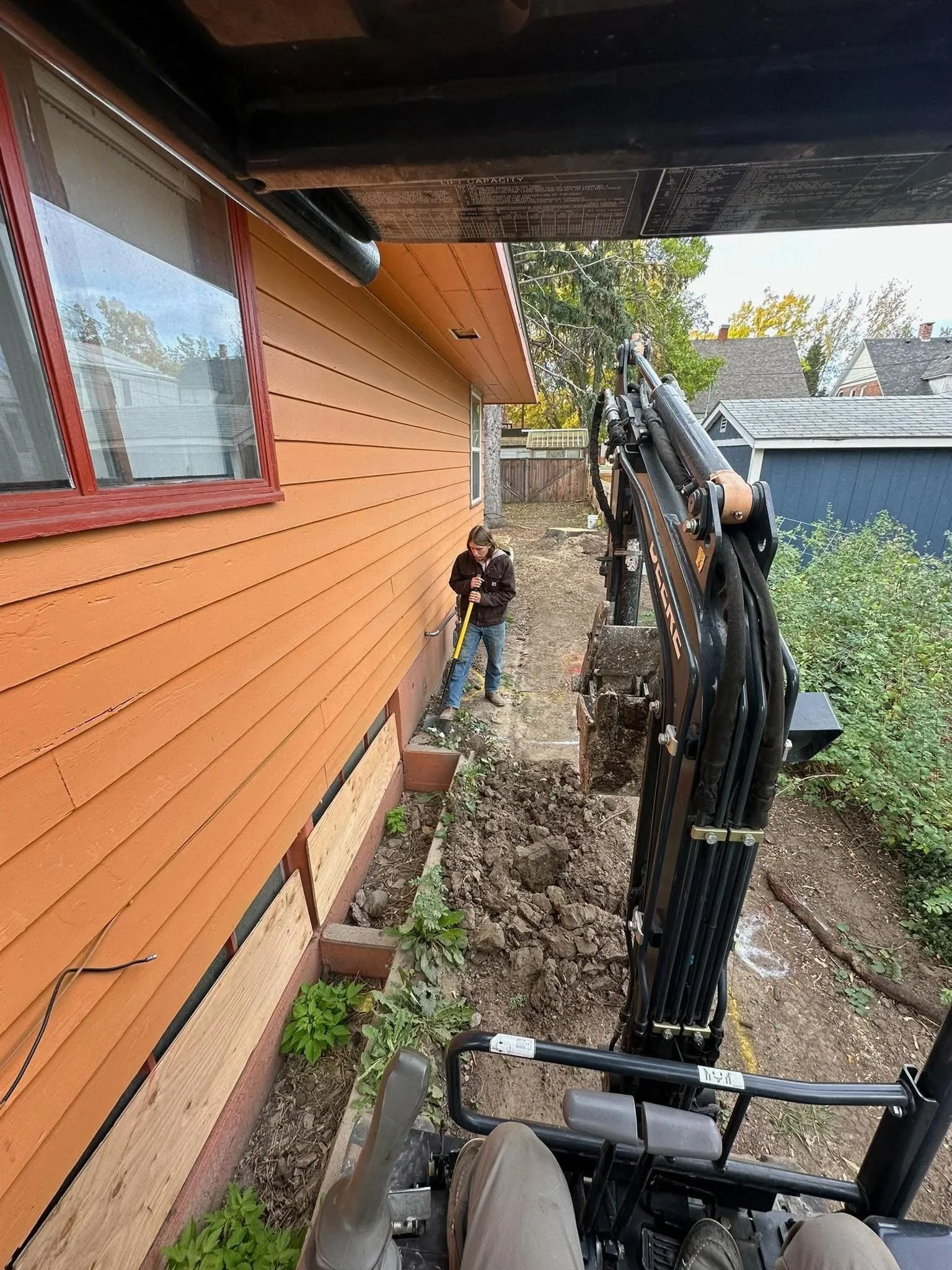 Person working with a construction excavator next to an orange house, digging a trench in the backyard.