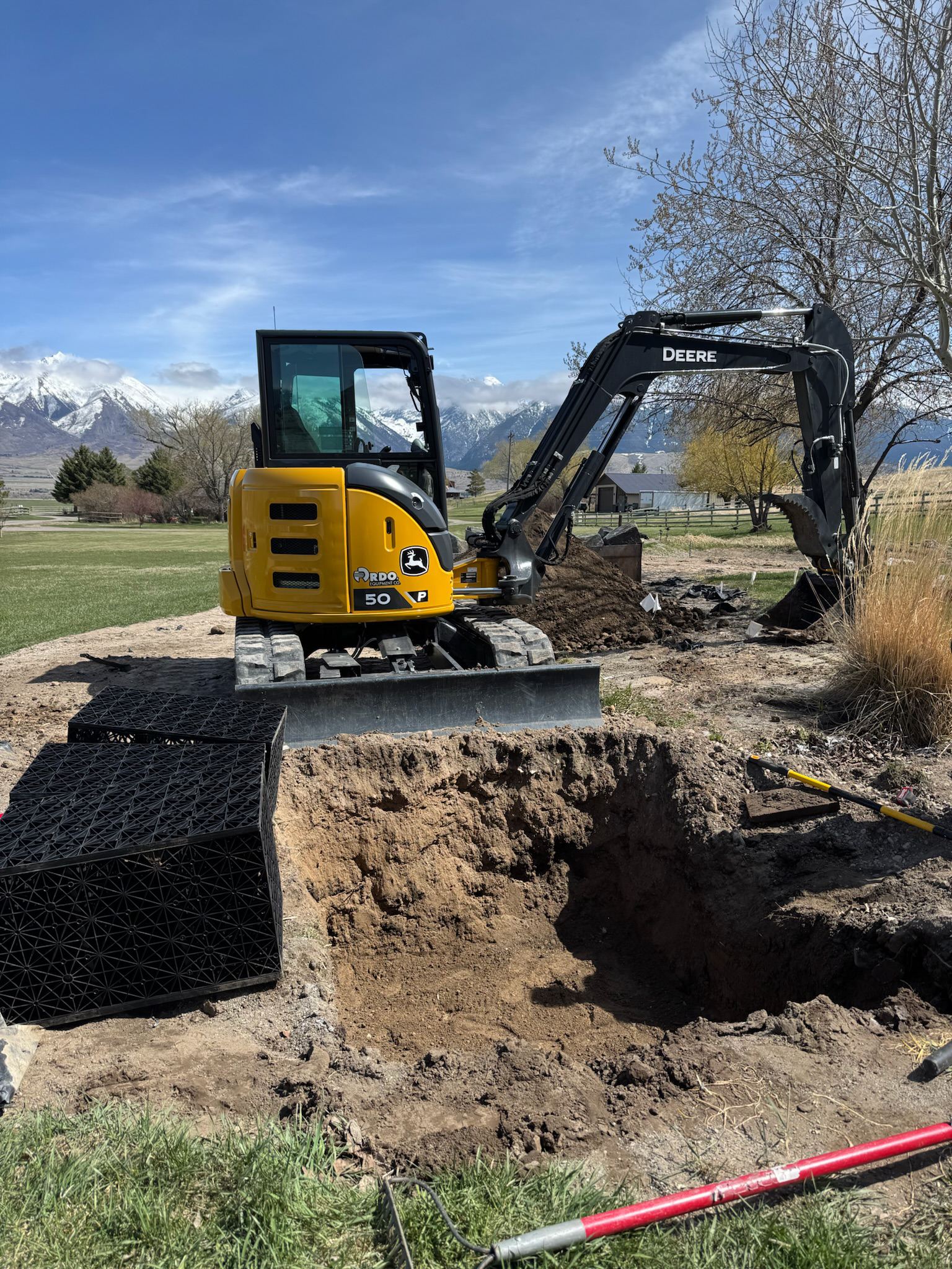 A small John Deere excavator digging a hole in the ground outdoors with mountains and blue sky in the background.