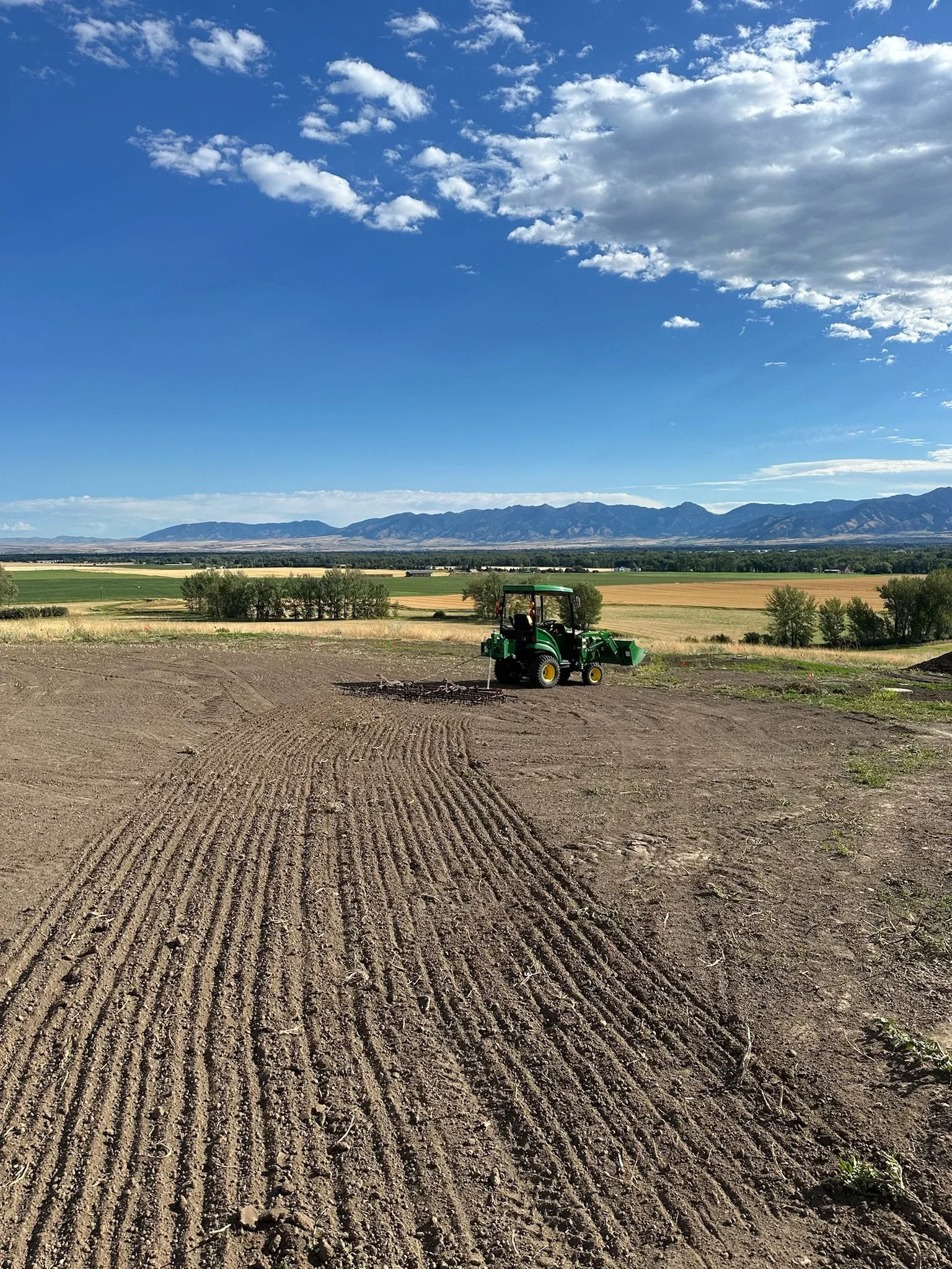 A tractor working in a field under a mostly clear blue sky with some clouds, with mountains in the background.