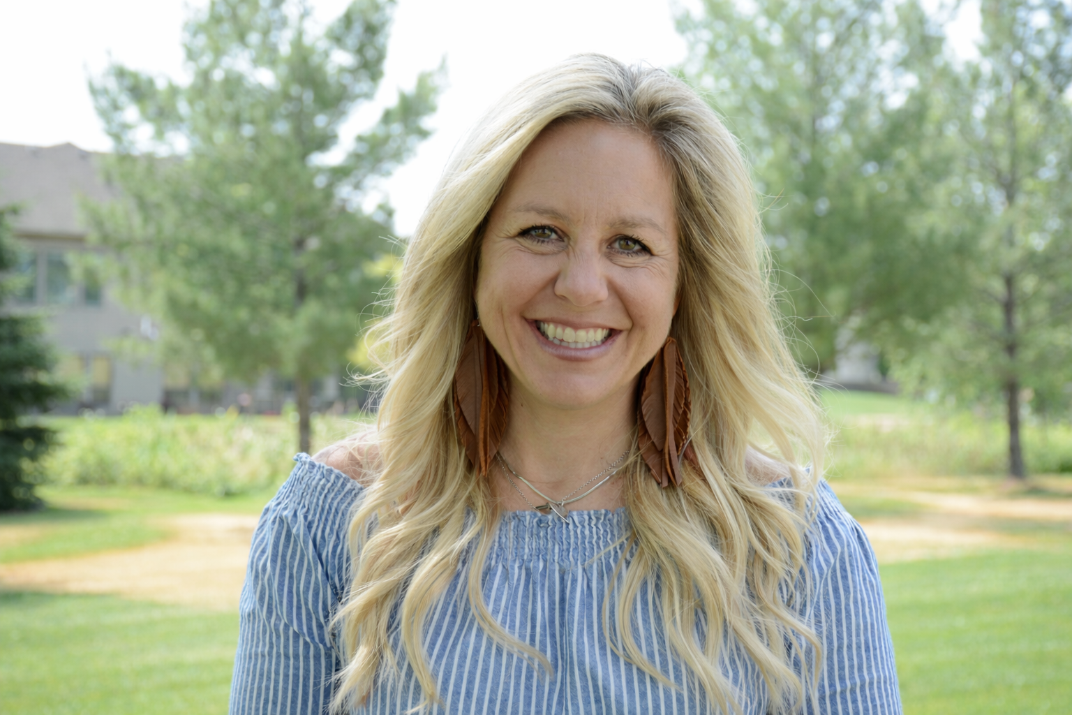 A woman with long blonde hair smiling outdoors in a park with trees and greenery in the background.