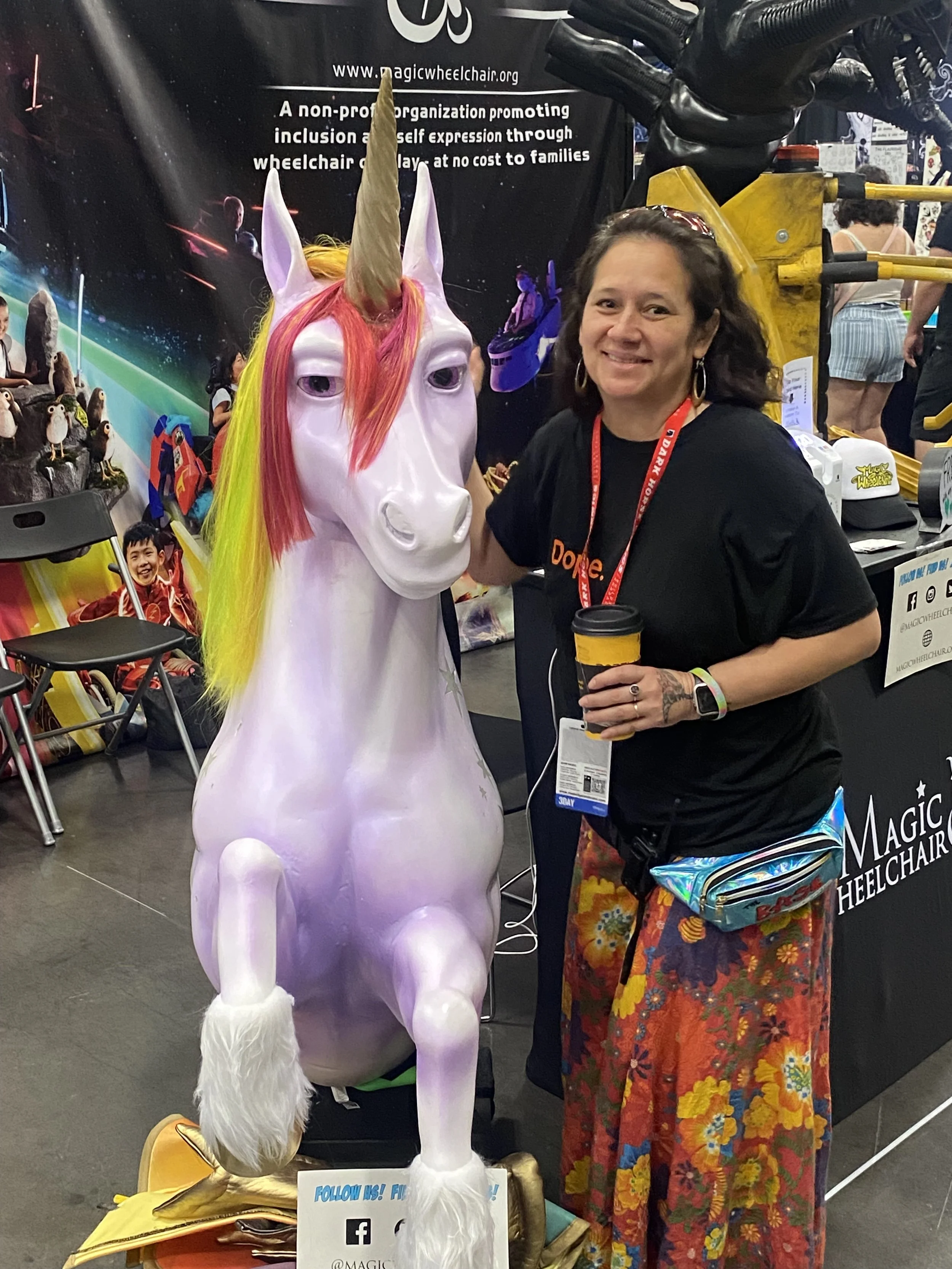 A woman smiling and holding a coffee cup stands next to a large, white unicorn mascot with a rainbow-colored mane and a golden horn at a convention booth.