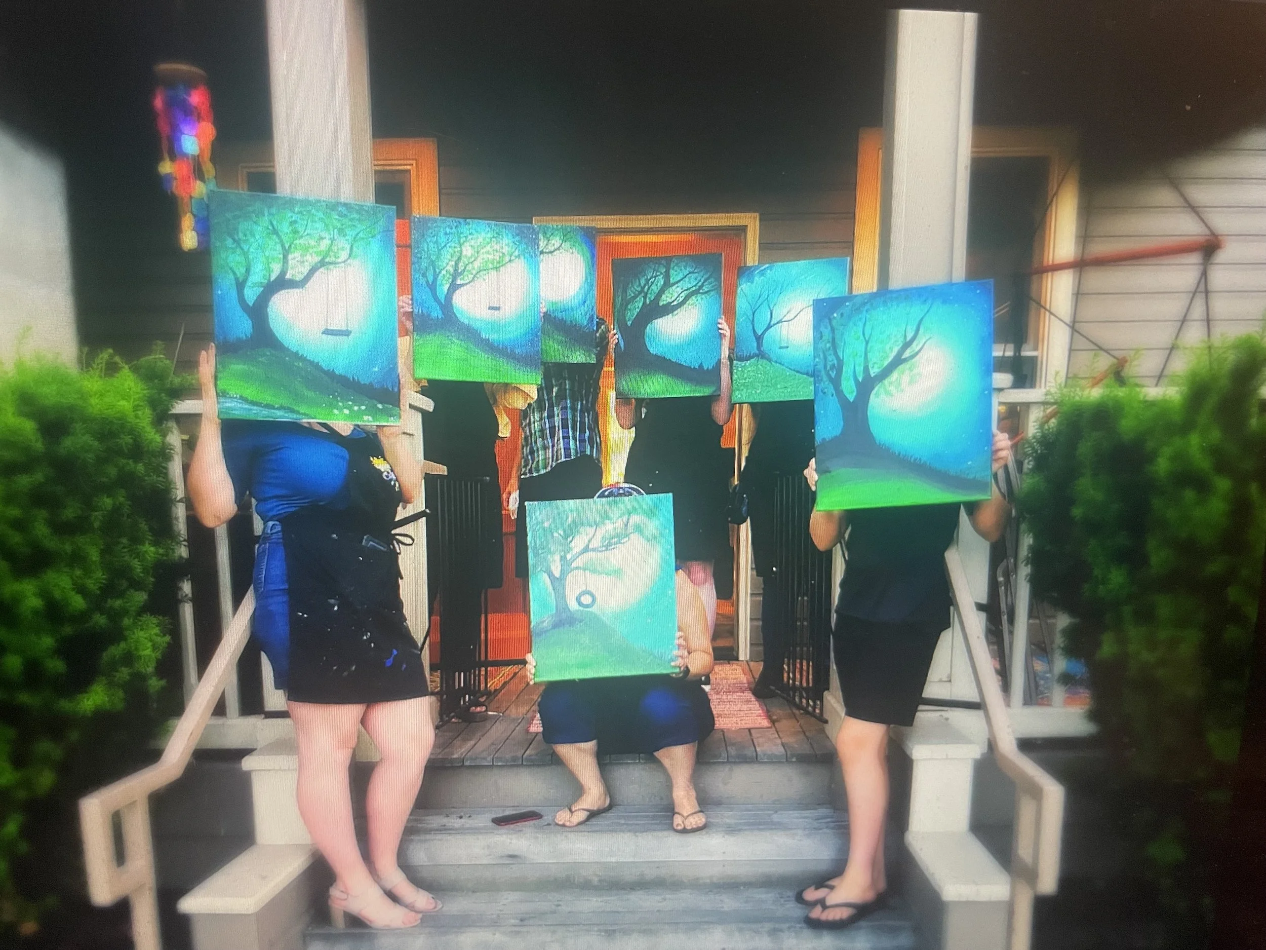 Group of four people holding paintings of trees with a moon, standing on a porch.