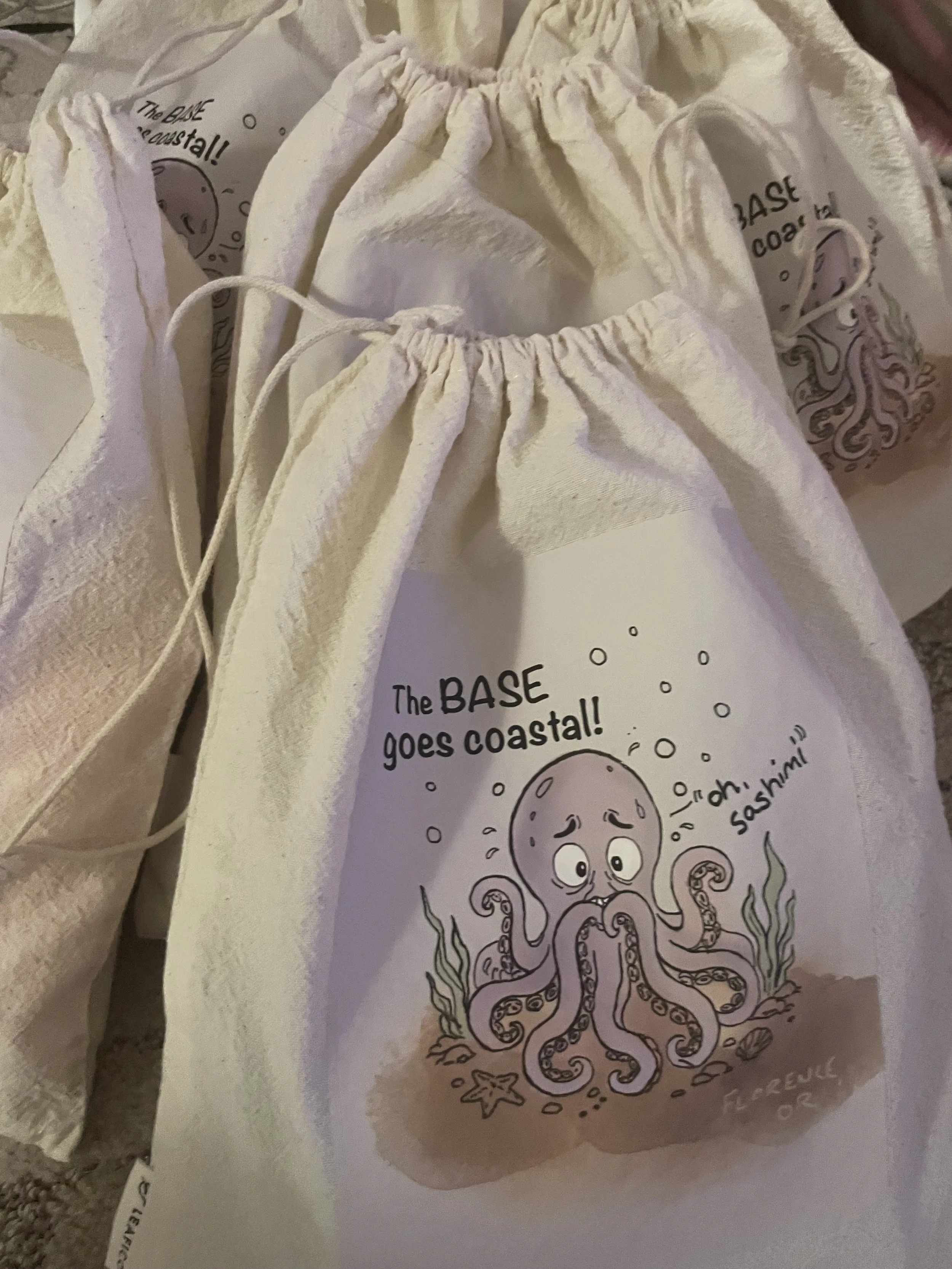 An illustration on a fabric bag of a cute octopus with a smiling face surrounded by bubbles and small starfish, with the text 'The BASE goes coastal!'