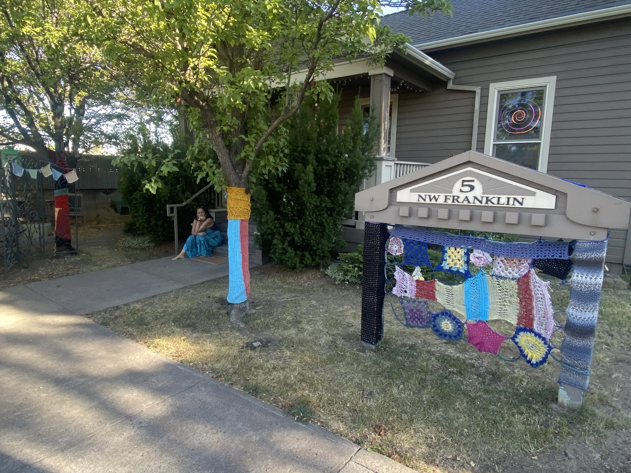 A front yard scene with a tree wrapped in colorful yarn, a woman sitting on a bench, and a house with a window displaying a neon-colored spiral. There are crocheted blankets hanging on a fence and tight-knit decorations on a blue table. The house has a gray exterior, and the street has a sidewalk and grassy area.