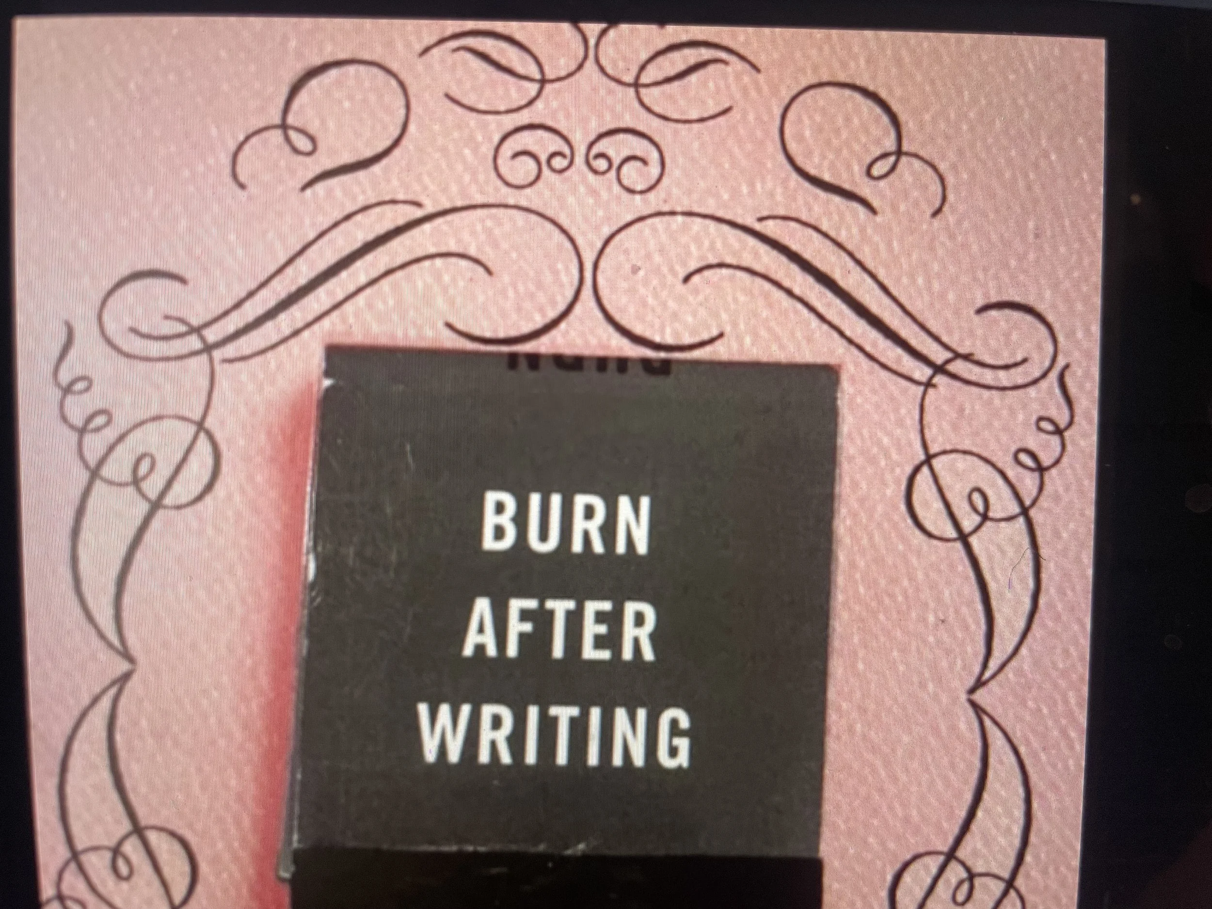 Close-up of a pink greeting card with black swirling designs. The card features a central black rectangle with white text that reads, "BURN AFTER WRITING."