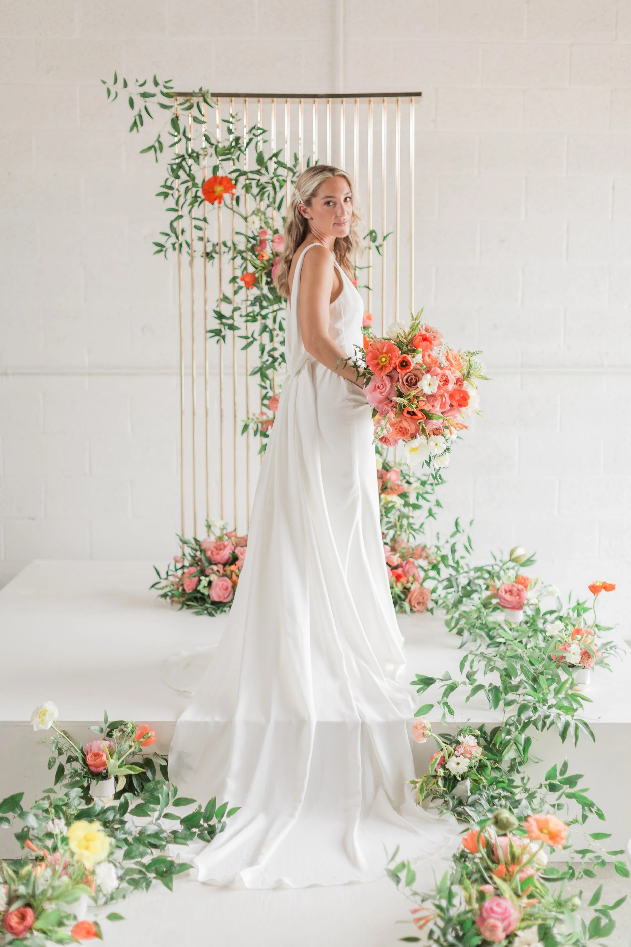 A woman in a white satin gown holding a large bouquet of pink and peach flowers in a decorated floral studio.