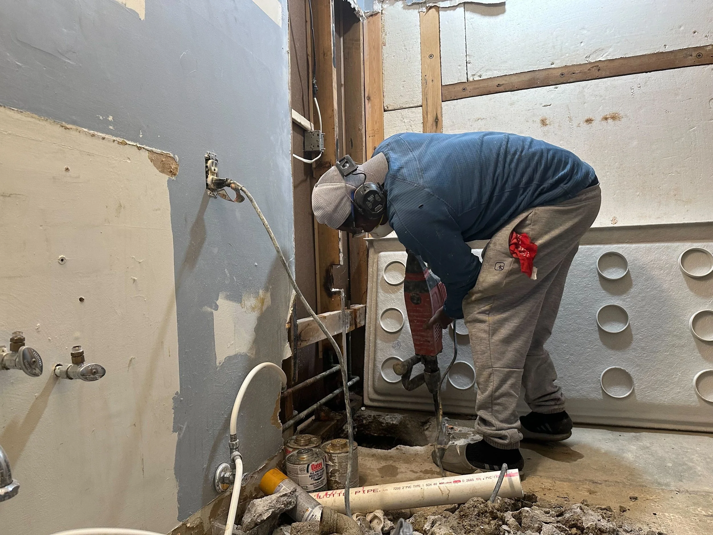 A construction worker wearing a gray cap, blue shirt, gray pants, and black shoes is using a jackhammer to break the concrete floor in a room under renovation.