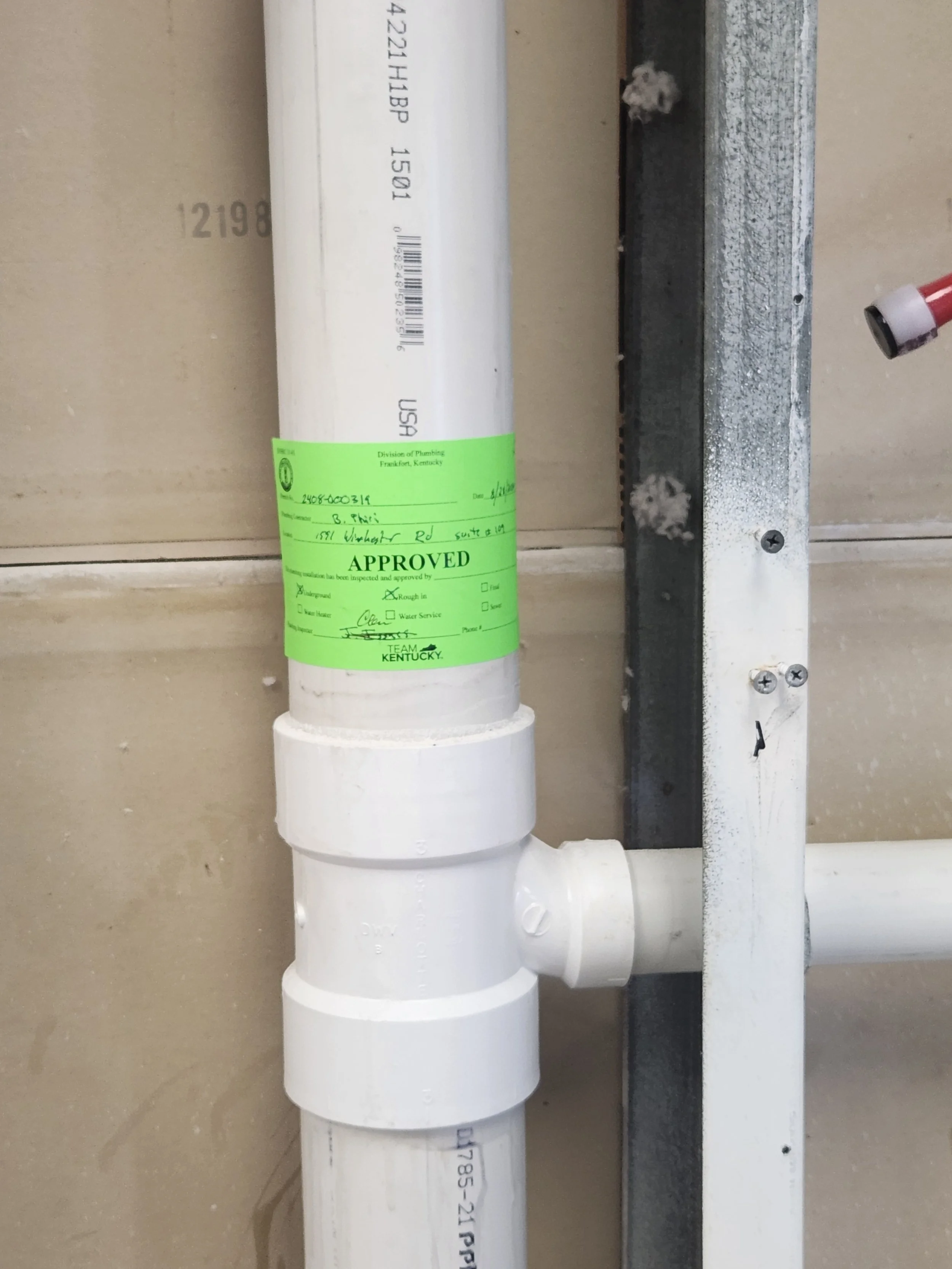 Plumbing pipe with an approval sticker attached, mounted on a wall with metal bracket, nearby are drywall screws and drywall surface.