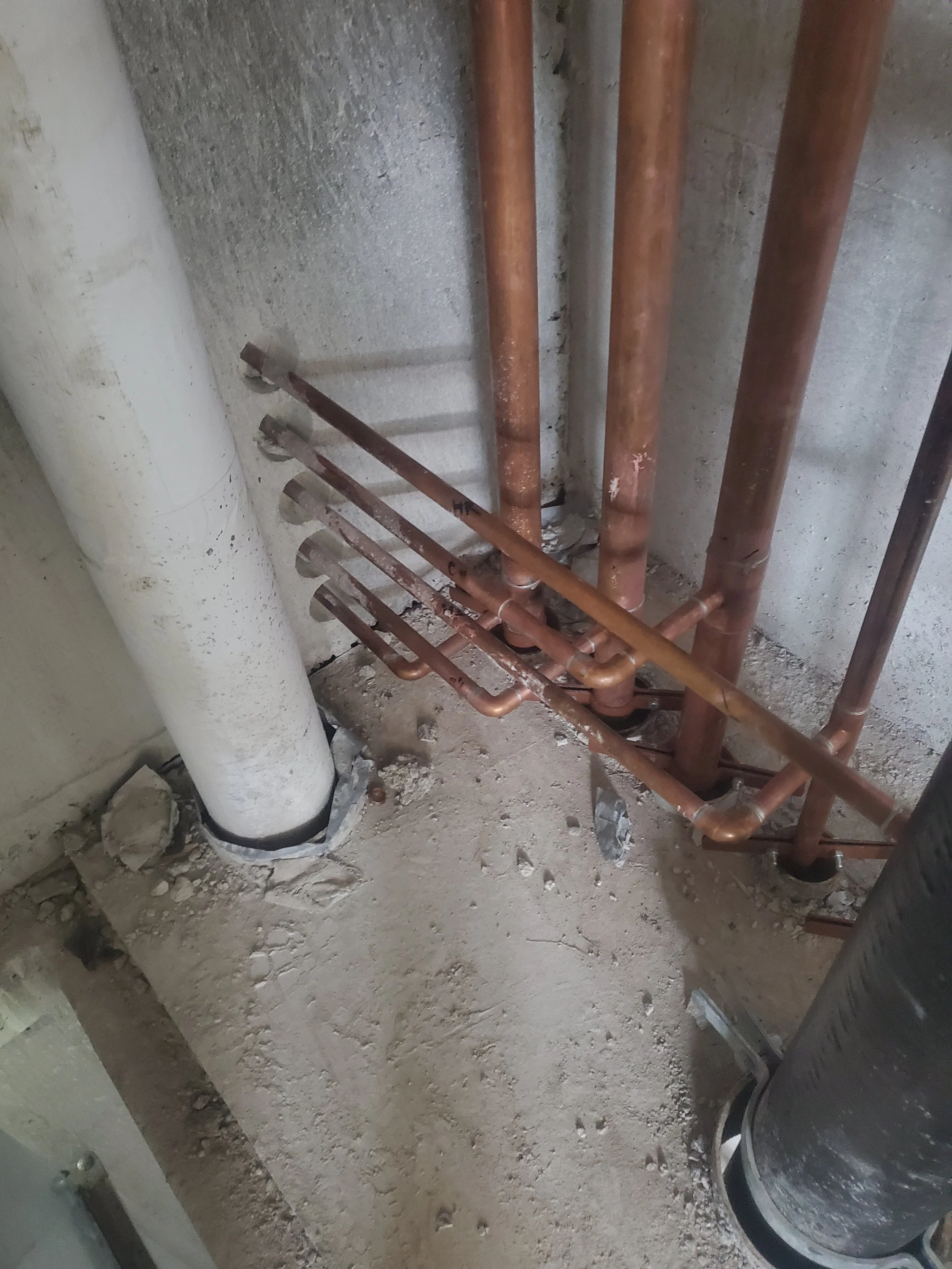 Close-up view of copper plumbing pipes installed on a concrete floor near a wall, with a large white pipe and a black vent pipe nearby, in a construction or renovation setting.