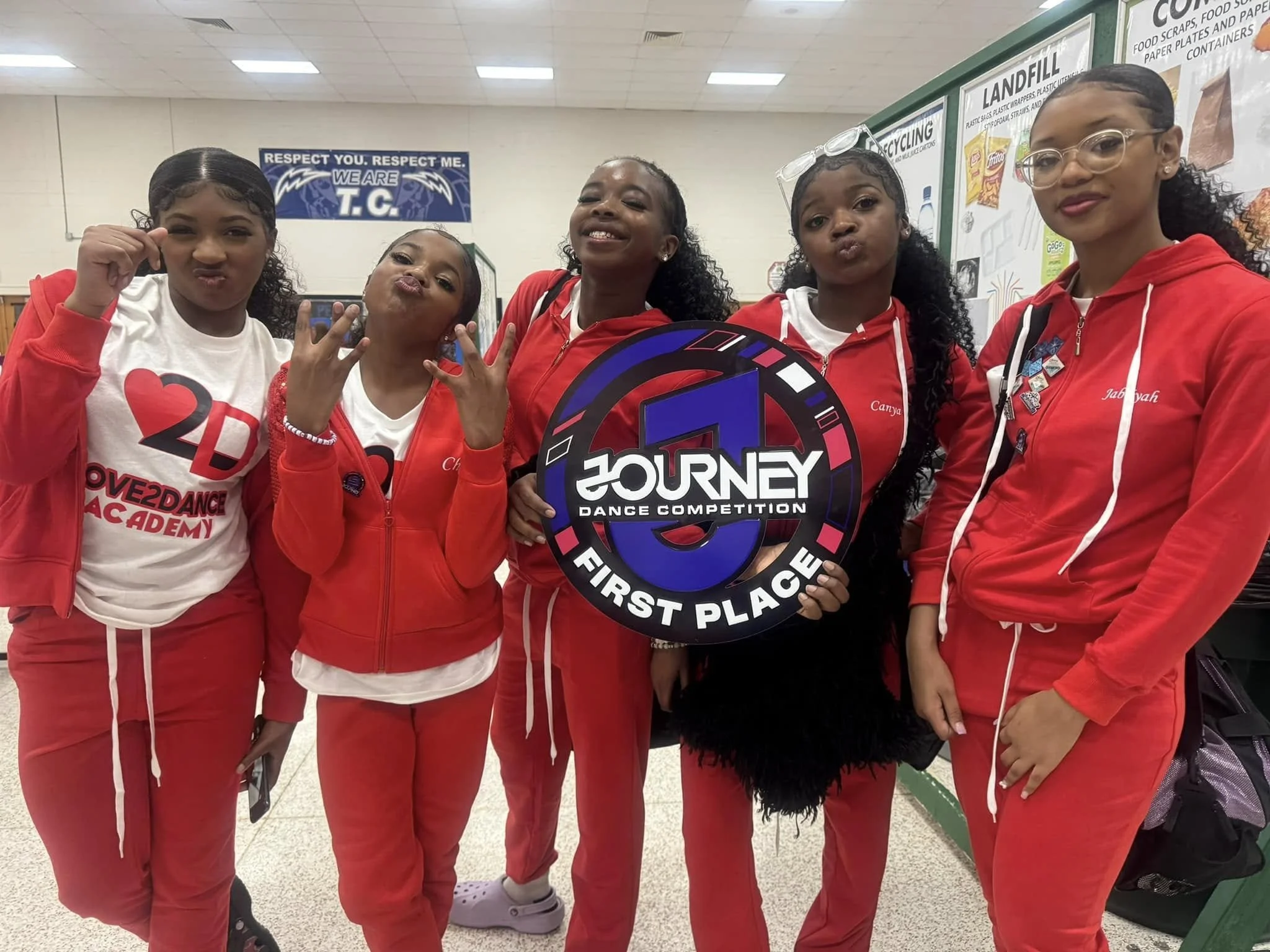 Five girls wearing red and white dance team outfits standing together indoors, holding a first place sign for a dance competition, smiling and making hand gestures.