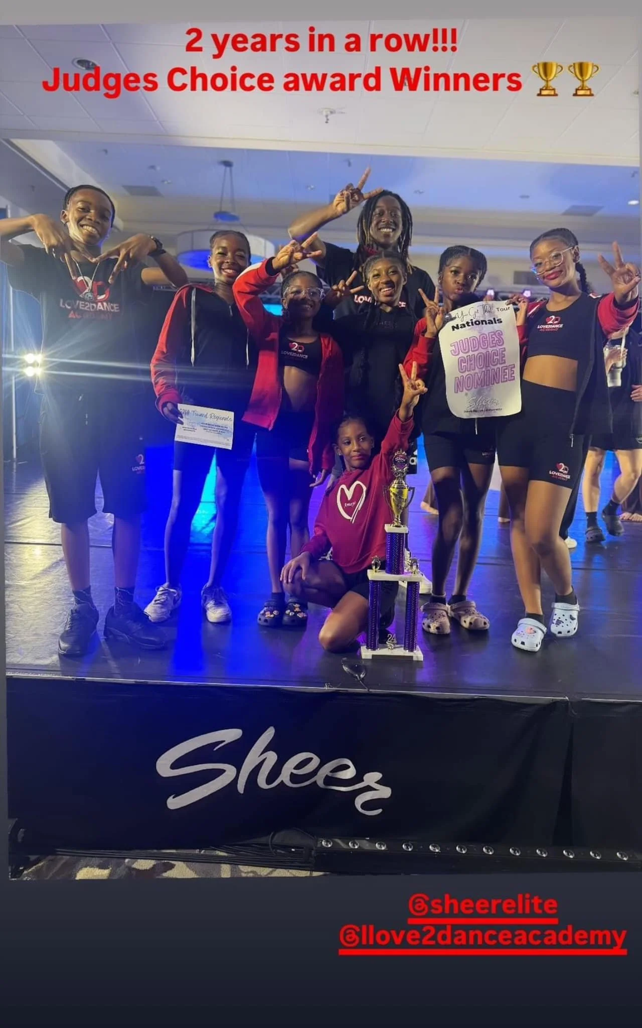 Group of young dancers on stage holding trophies and certificates, celebrating their awards, with a sign reading 'Judges Choice Nominee' and a banner with 'Sheer' visible at the bottom, indicating they are winners of a dance competition.