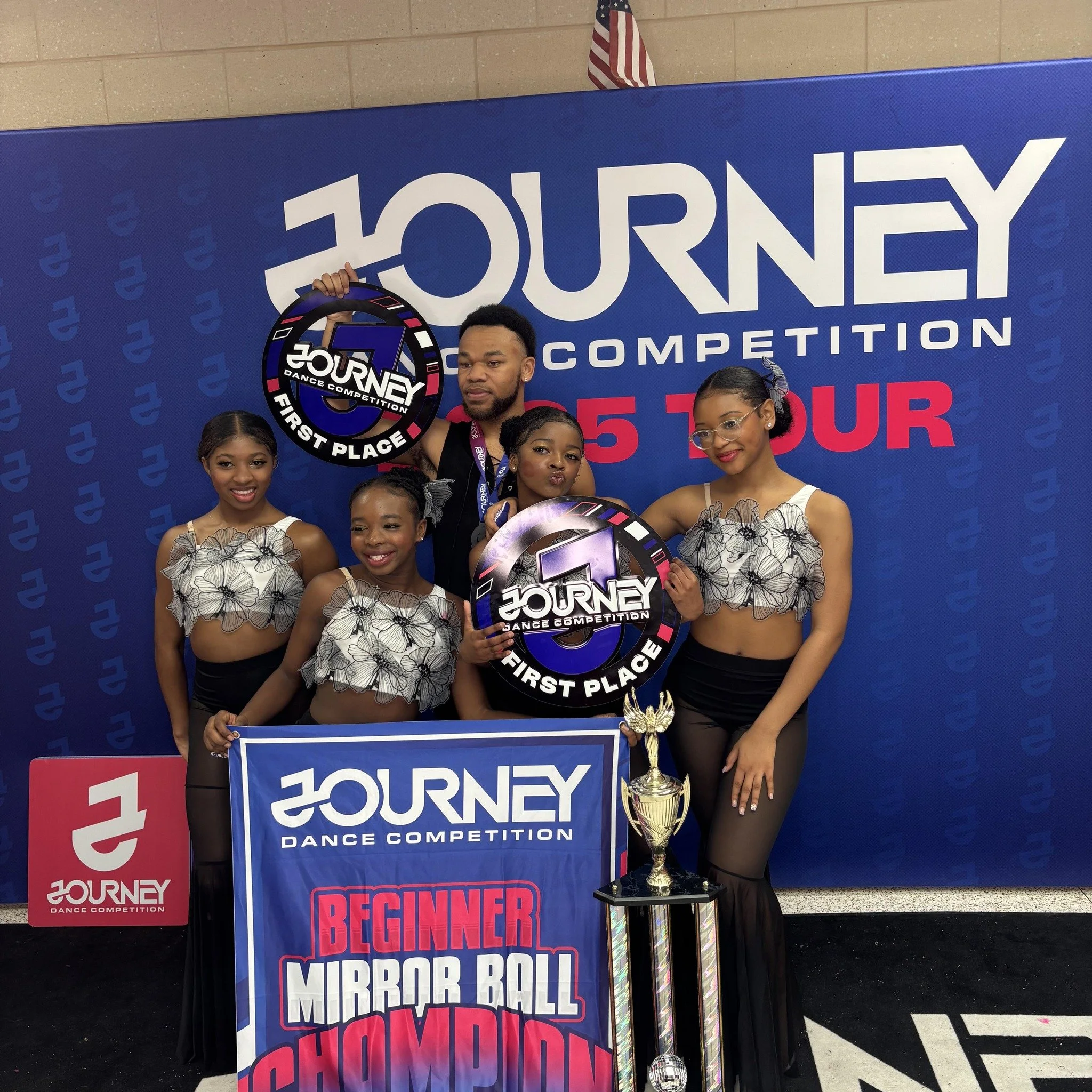 A group of five dancers at a dance competition, holding trophies and signs that say "First Place," standing in front of a blue backdrop with the text "Journey Dance Competition" and "Beginner Mirror Ball Showdown."