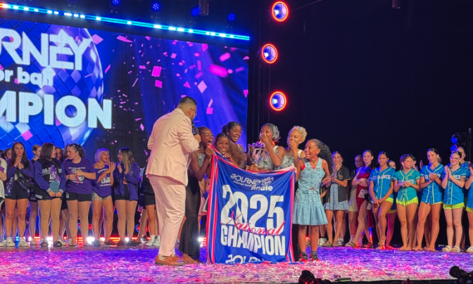 Group of cheerleaders on stage celebrating championship victory, holding a large blue banner with '2025 Champion' written on it, surrounded by confetti and cheering.