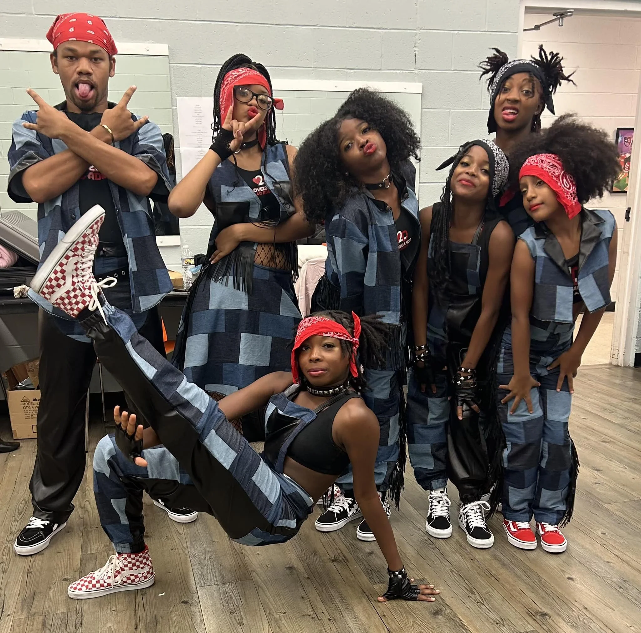 A group of seven young adults dressed in matching blue and black checkered outfits with red bandanas, standing in a room with white walls and wooden flooring, posing for a fun photo with various expressive gestures.