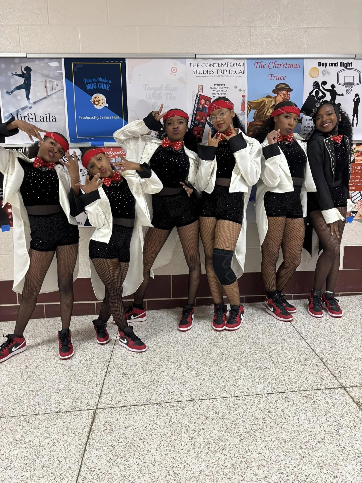 Six young girls dressed in coordinated dance outfits pose in a school hallway in front of a bulletin board, wearing black and white clothing with red accessories and sneakers.