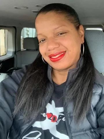 Woman with long black hair, red lipstick, and hoop earrings smiling inside a vehicle.