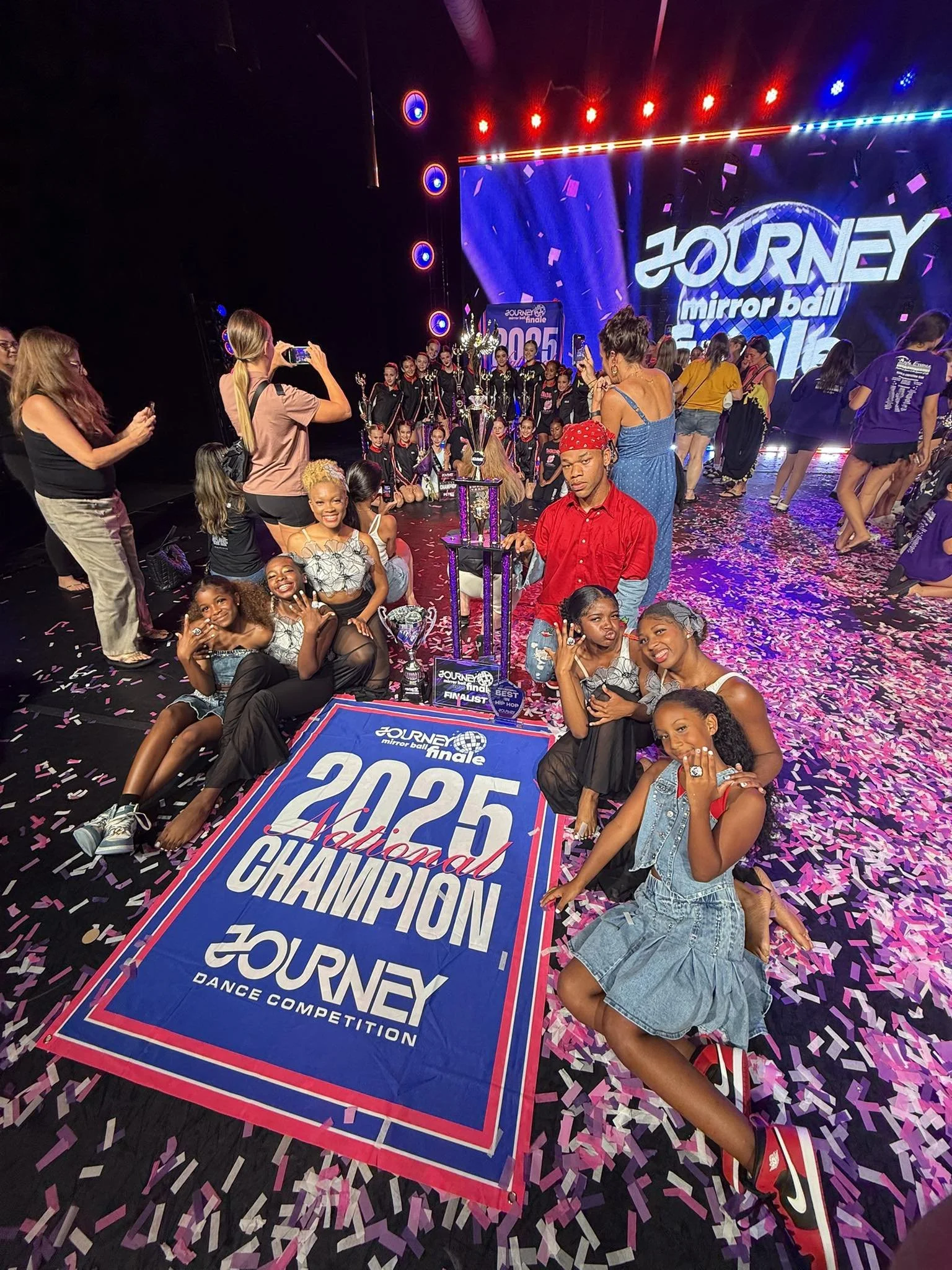 Group of young dancers celebrating their victory at the 2025 Journey dance competition. They are posing with trophies and a banner on a stage decorated with pink and purple confetti, with a large screen displaying the event's name and year.