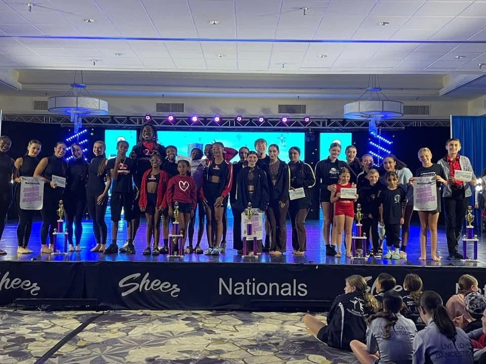 Group of young female cheerleaders and dancers on stage holding trophies and certificates at a cheerleading competition, with an audience watching.