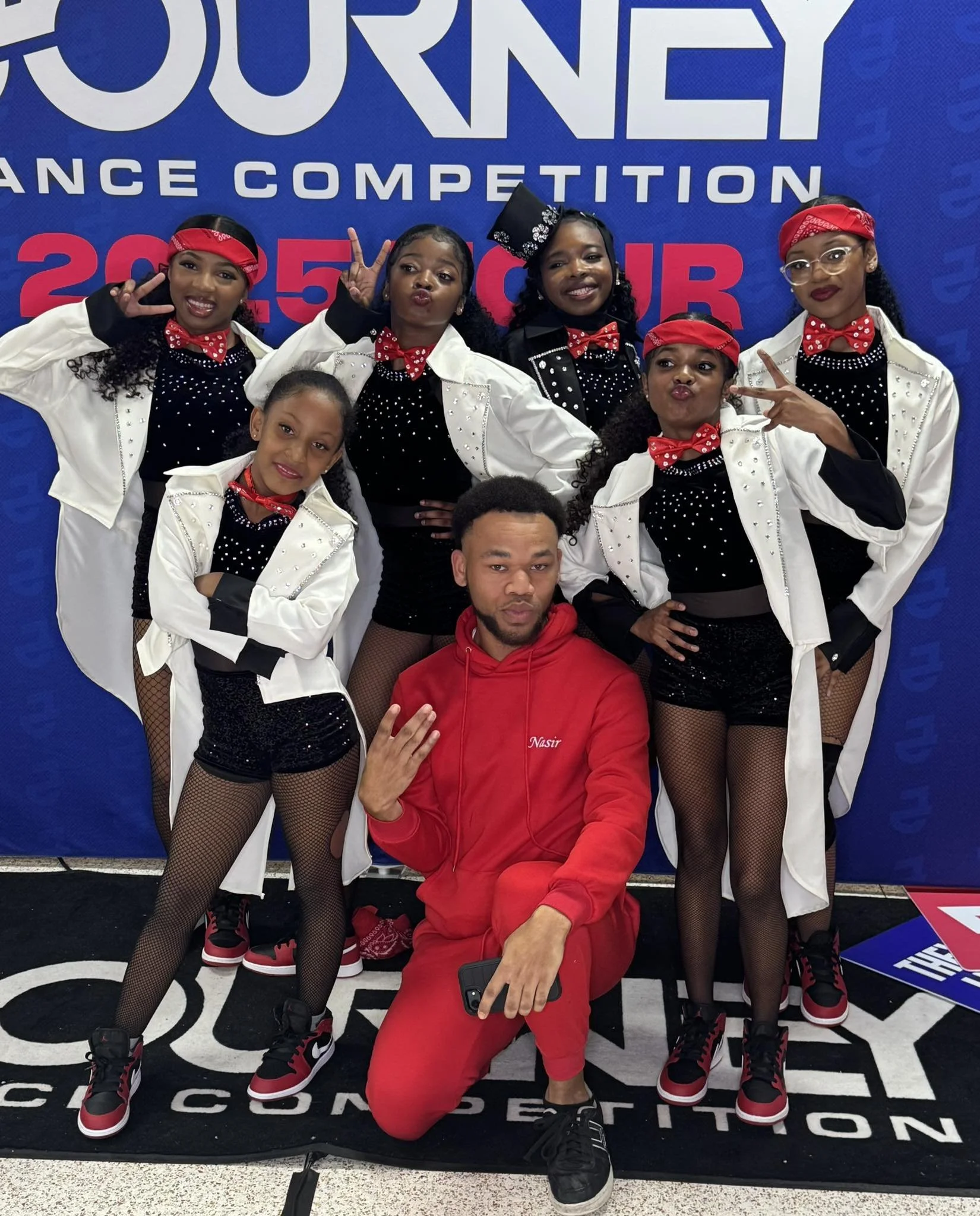 Group of young dancers in black sequined shorts, fishnet tights, white jackets, red bandanas, and black and red sneakers, posing with their coach in front of a blue backdrop at a dance competition.
