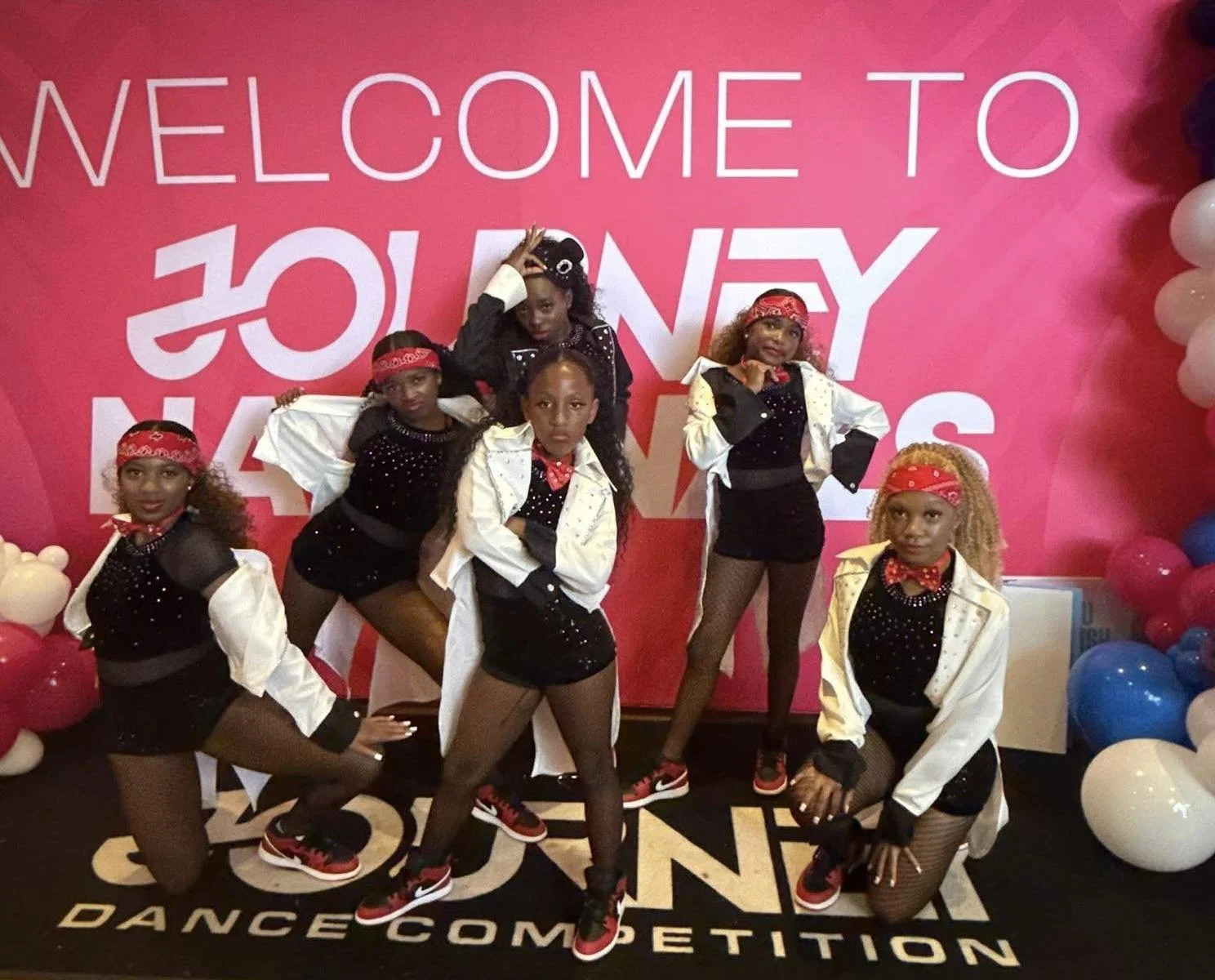 Group of young dancers in coordinated costumes posing in front of a pink backdrop with white and red balloons at a dance competition event.