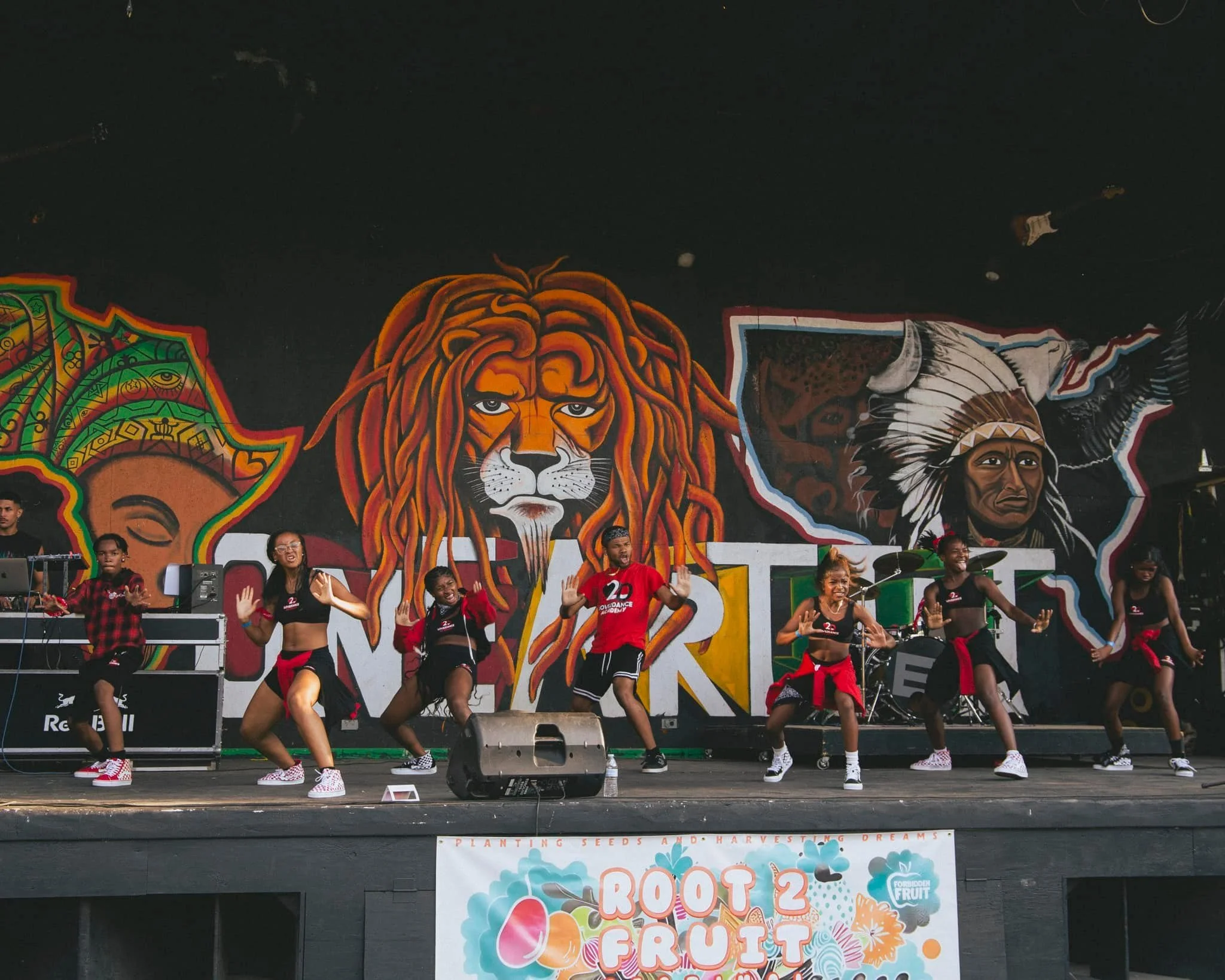 A group of eight dancers performing on stage in front of a colorful mural including a lion and a Native American chief. The dancers are wearing black and red outfits.