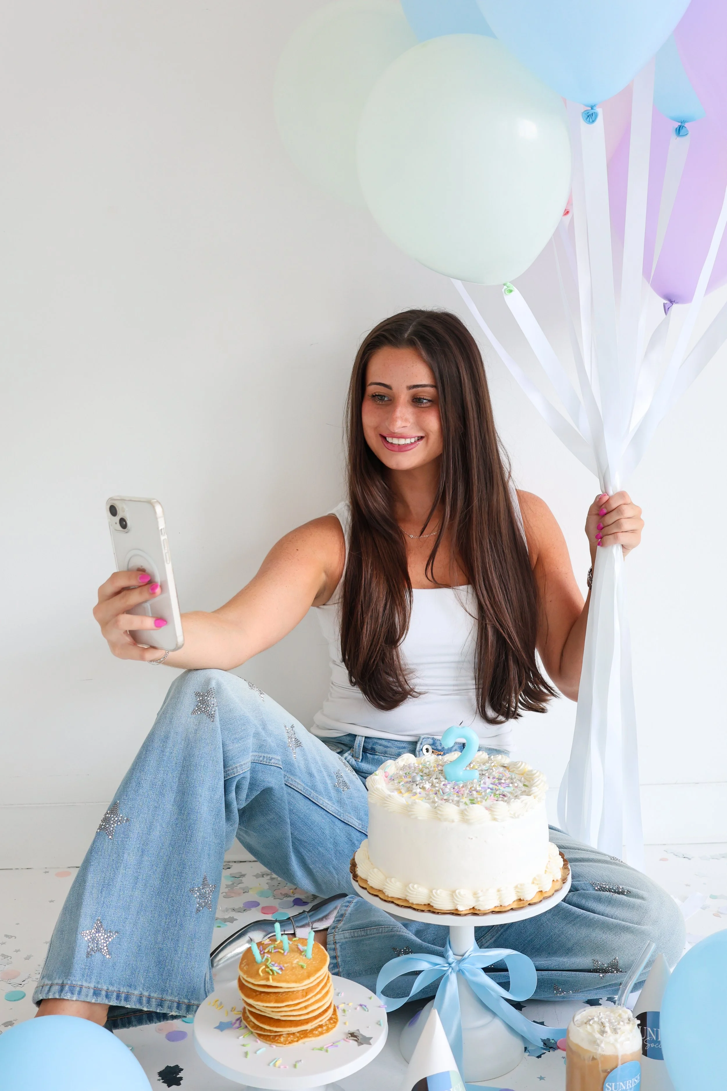 A young woman with long brown hair taking a selfie with her phone, celebrating a birthday with balloons, cake, cupcakes, and decorations.