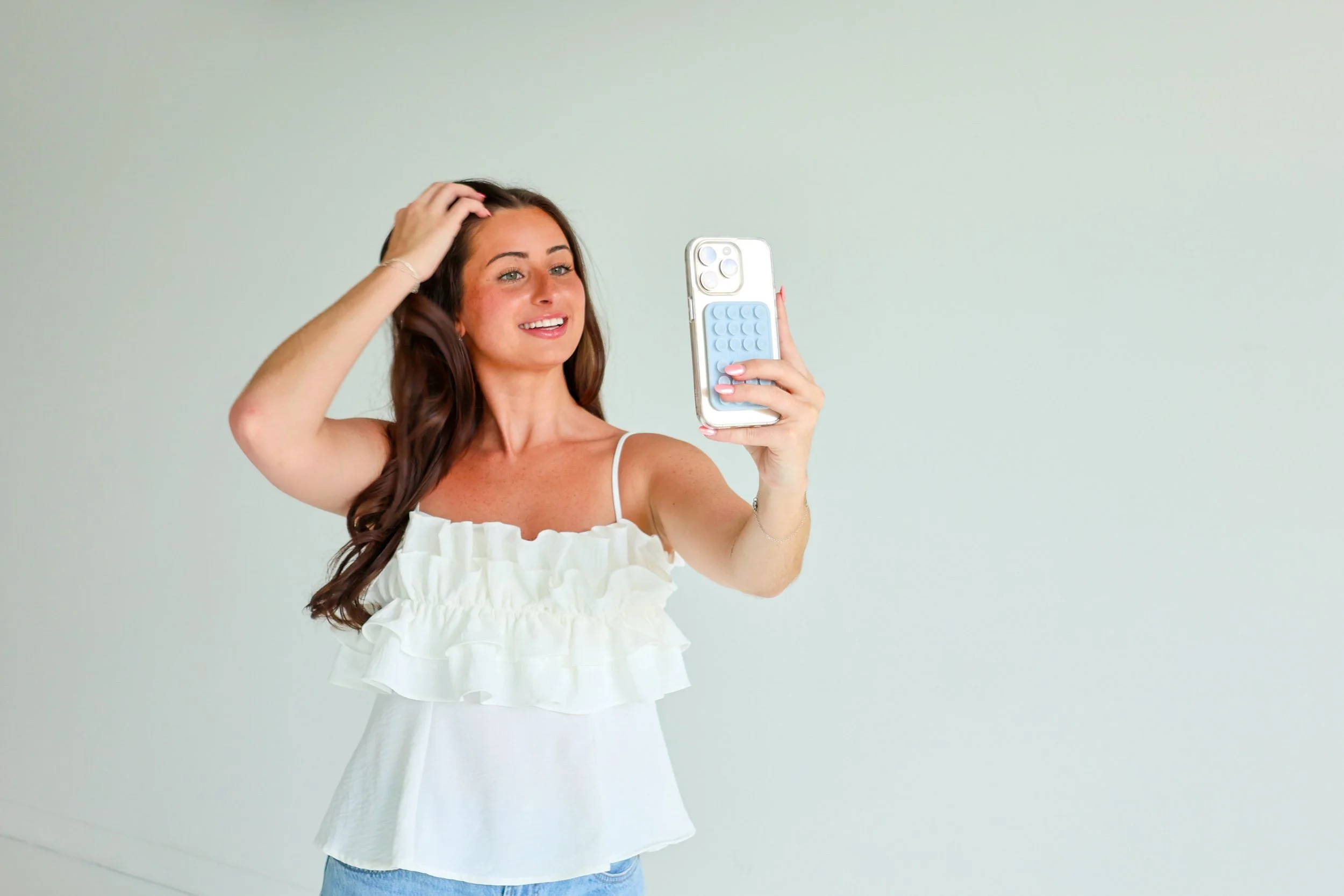A woman with long brown hair, wearing a white ruffled top, is taking a selfie with her smartphone, which has a blue phone grip. She is smiling and touching her hair with her left hand. She is standing against a plain light-colored wall.