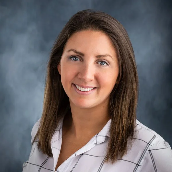 A woman with shoulder-length brown hair and blue eyes smiling at the camera, wearing a white shirt with thin black stripes against a neutral background.