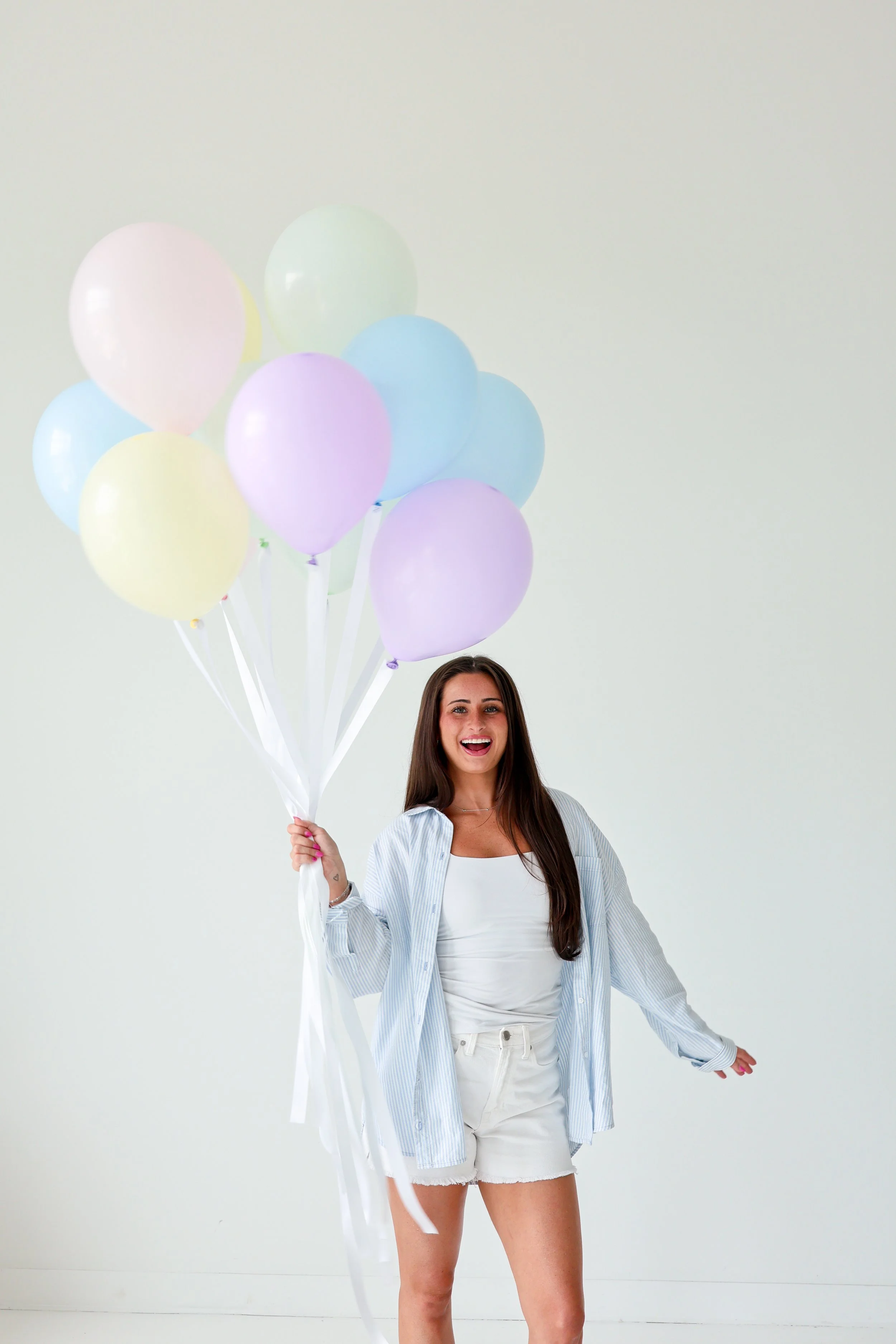 A young woman smiling and holding a bunch of pastel-colored balloons while standing against a plain white wall.
