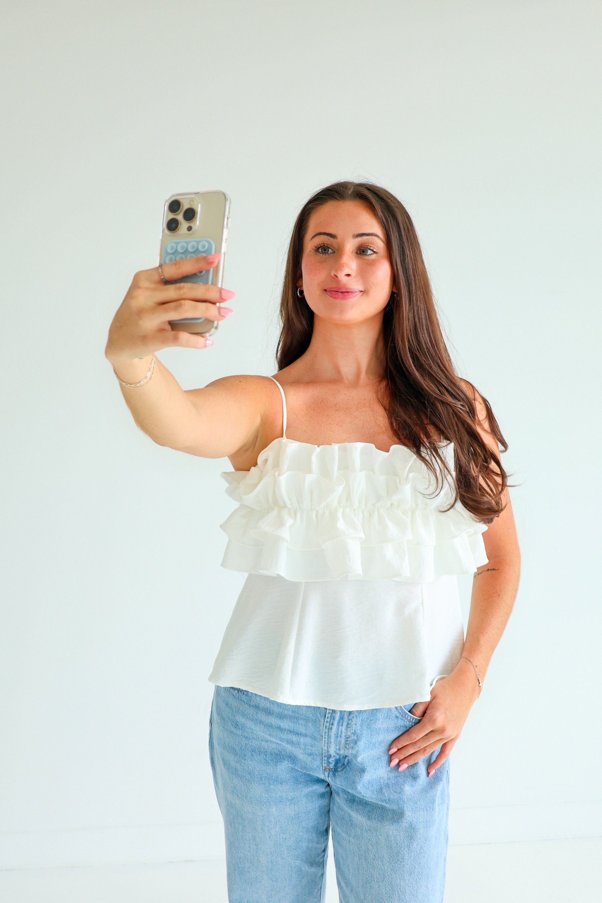 Young woman with long brown hair taking a selfie with her smartphone, wearing a white ruffled sleeveless top and light blue jeans against a plain white background.