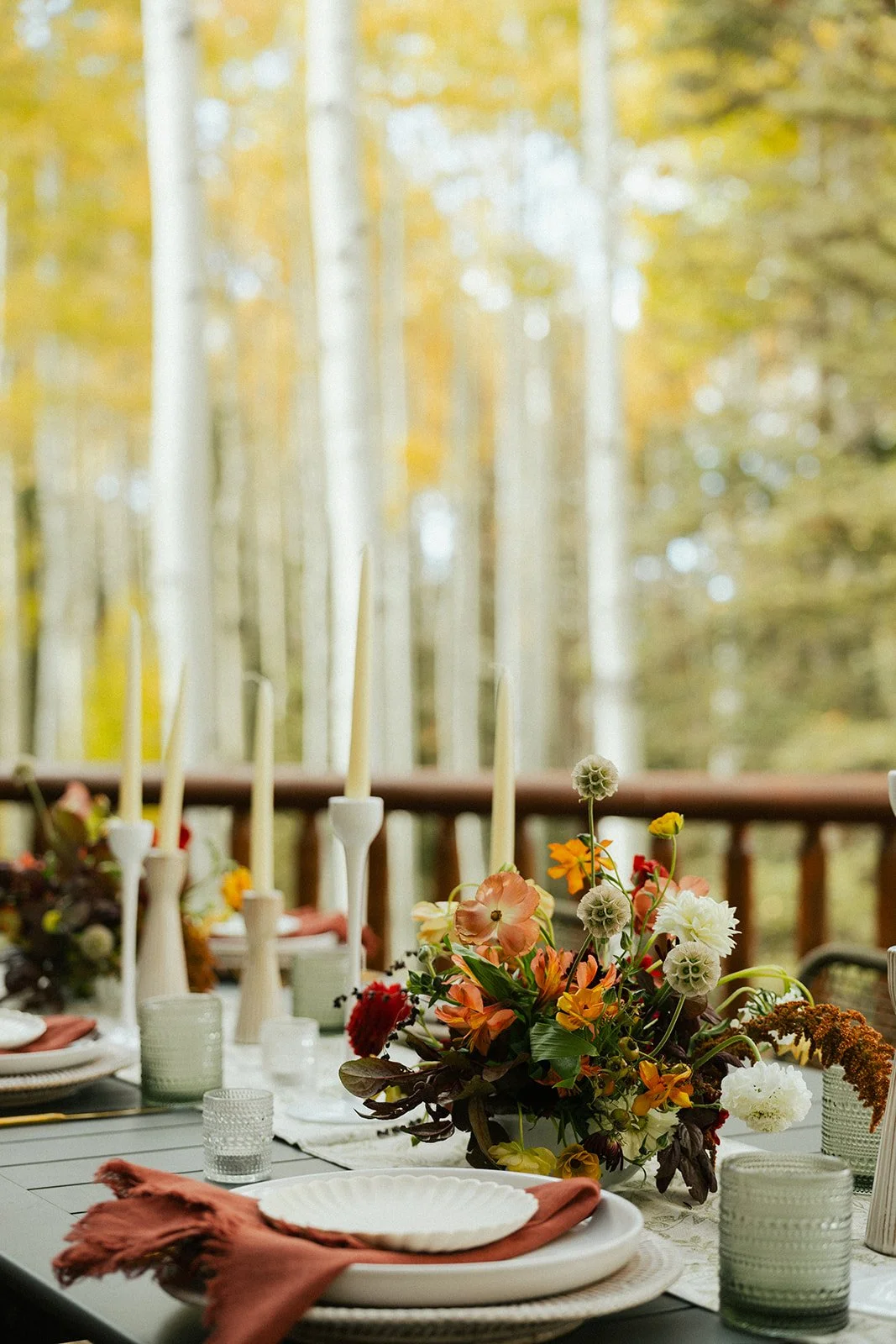 A decorated outdoor dining table with a floral centerpiece, candles, plates, glasses, and napkins set for a meal on a wooden deck, with a forest of Aspen trees in the background.