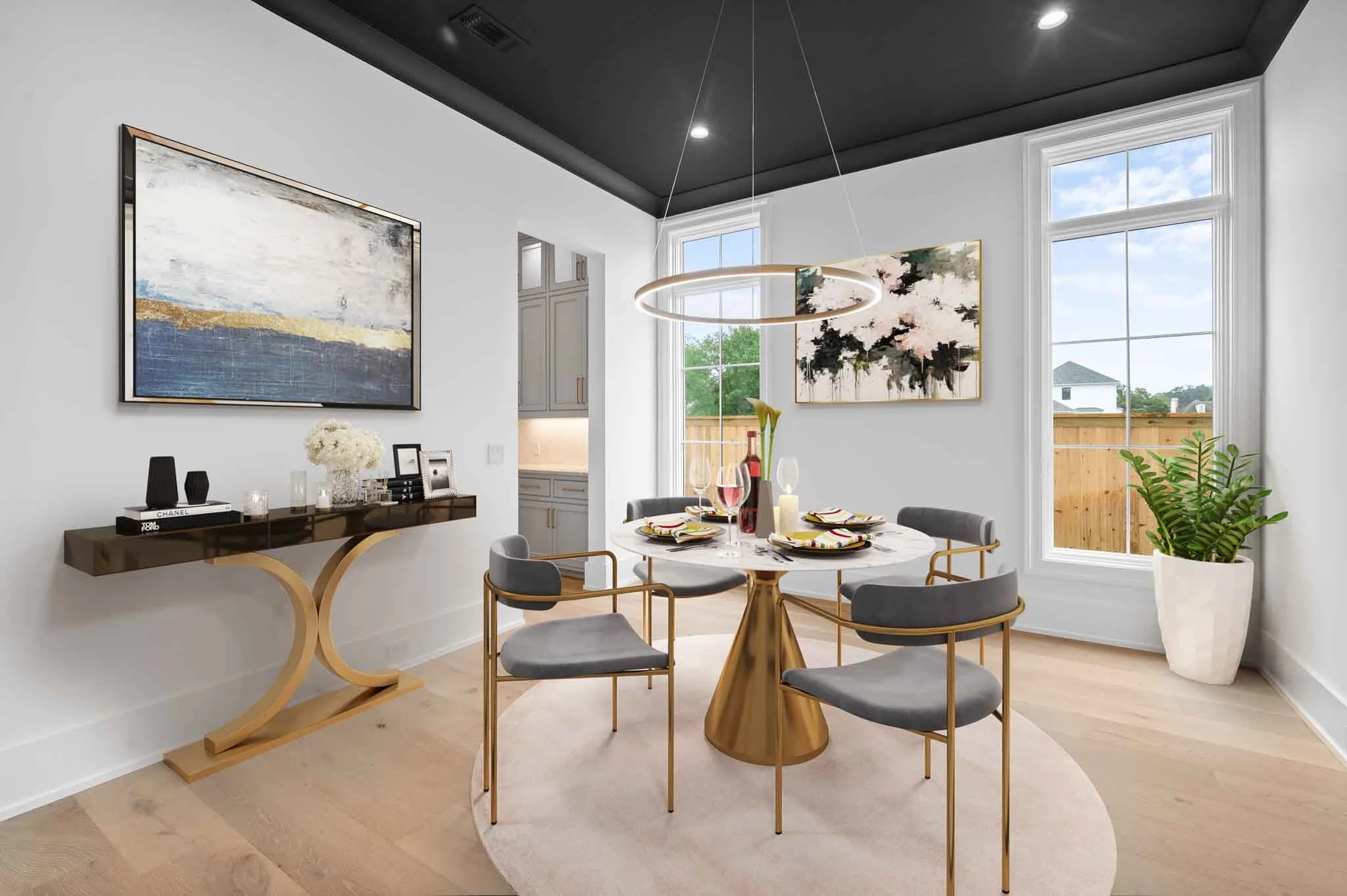 Modern dining room with a round table, gray chairs with gold frames, and contemporary artwork on the walls. Large windows allow natural light, and a potted plant is in the corner. A sleek console table is against the wall with decorative items.