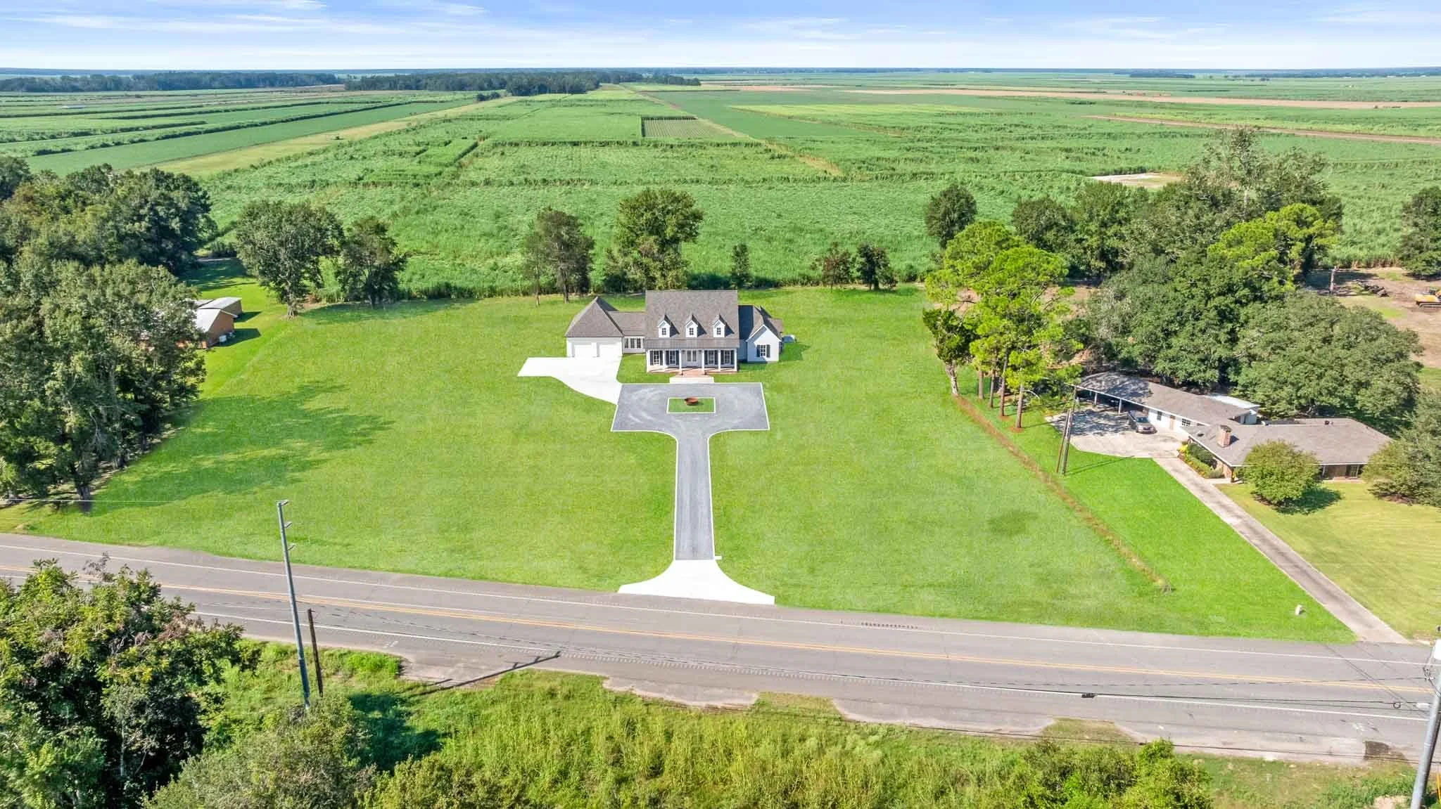 Aerial view of a large house with a gray roof, white exterior, and a front porch, situated on expansive green lawn. A paved driveway leads from the road to the house, with a small white area at the entrance. Surrounding the property are trees and open farmland with fields extending into the distance.