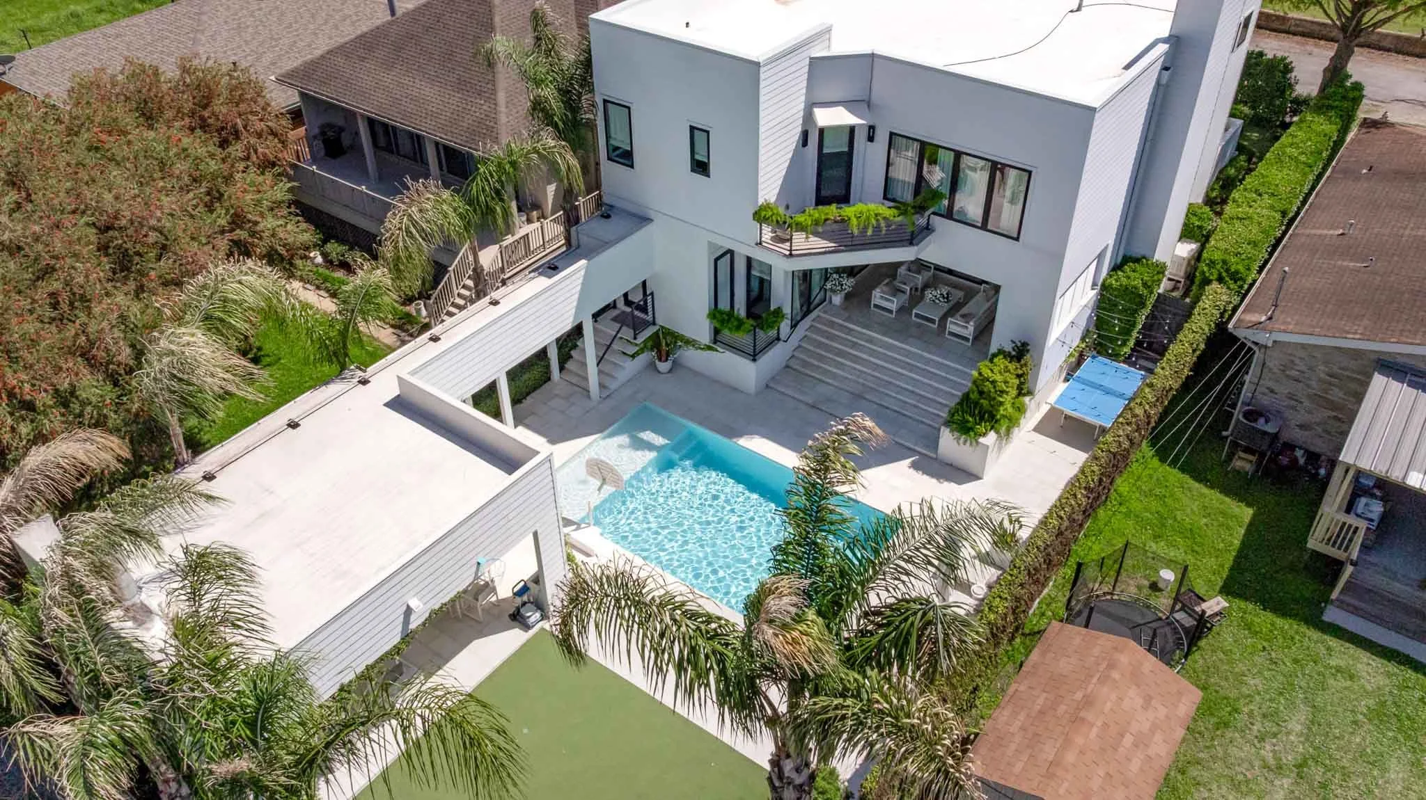 A modern white house with an outdoor pool in the backyard, surrounded by green foliage and neighboring houses.