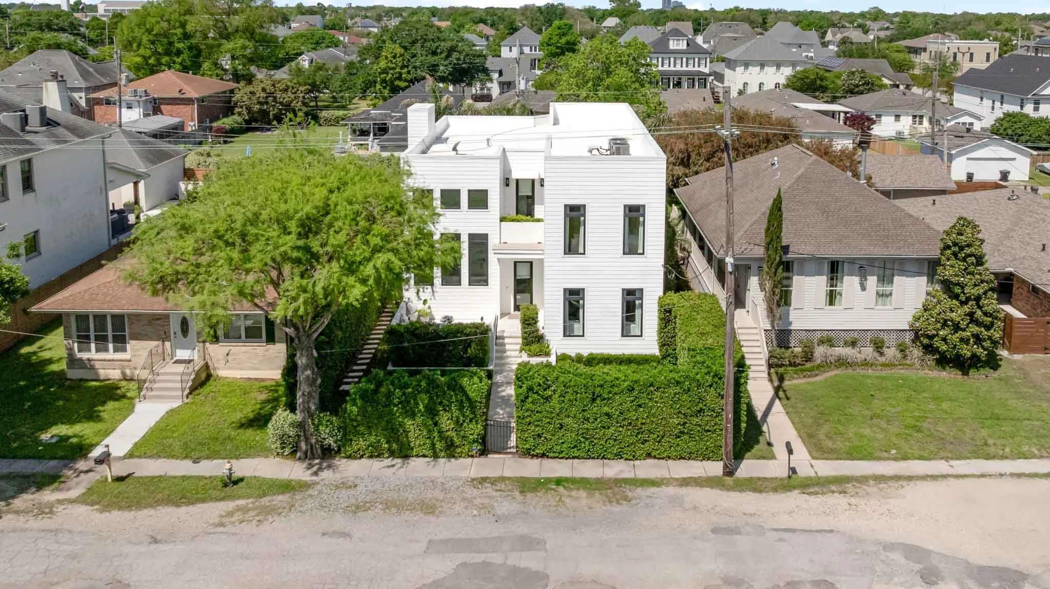 An aerial view of a residential neighborhood showing a modern white house in the center, with a mature tree and manicured bushes in front, and neighboring houses with different architectural styles and rooftops, surrounded by greenery.