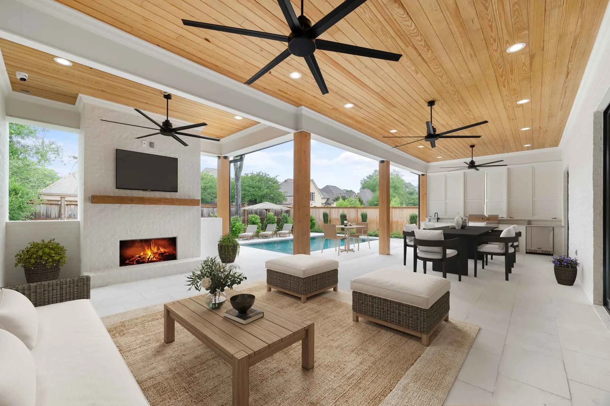 Covered backyard patio with ceiling fans, fireplace, television, outdoor seating area, dining table, kitchen area with cabinets and sink, swimming pool with poolside lounge chairs, and surrounding greenery.