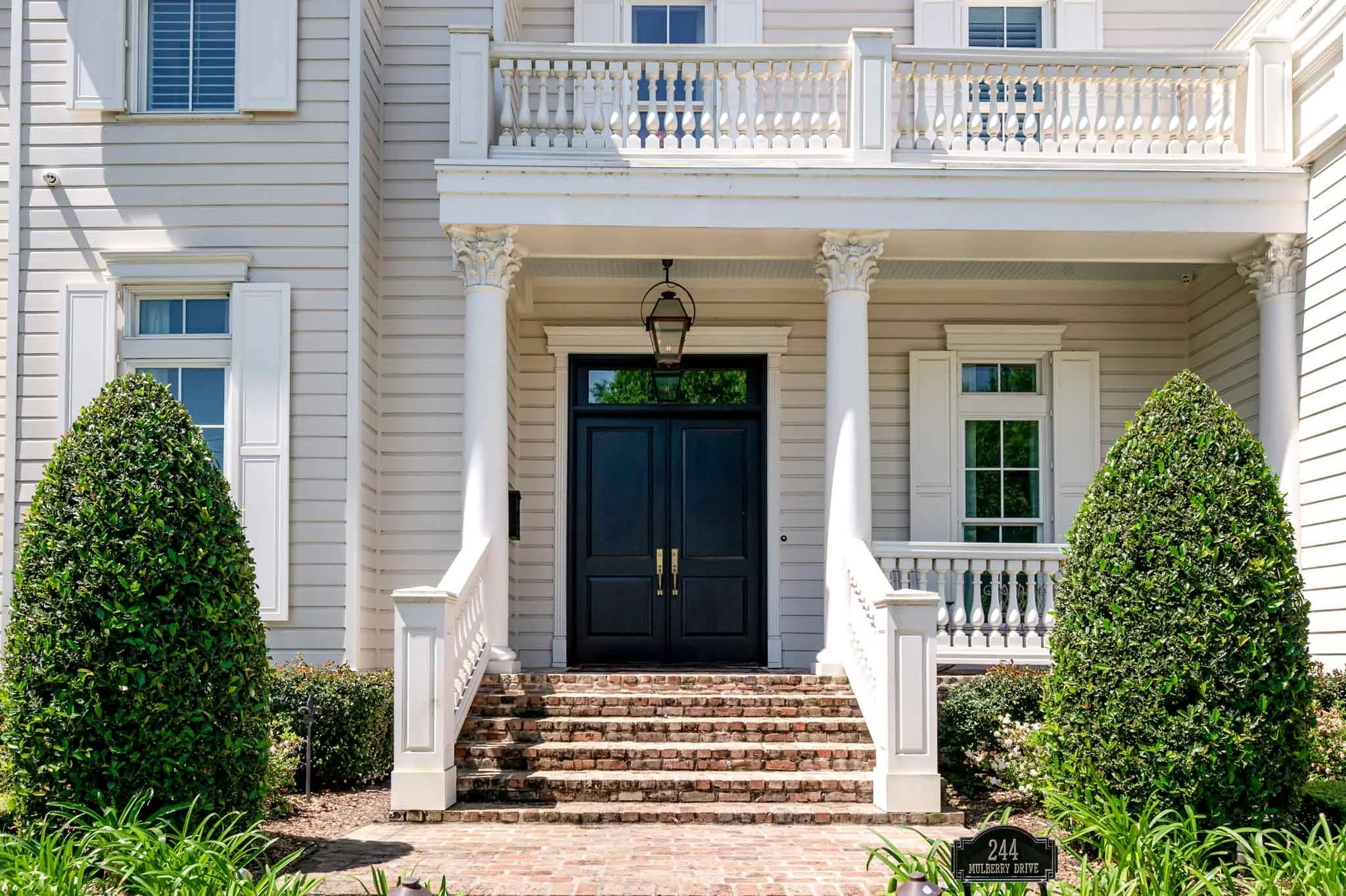 Front entrance of a white house with black double doors, brick steps, potted plants, and neatly trimmed bushes.