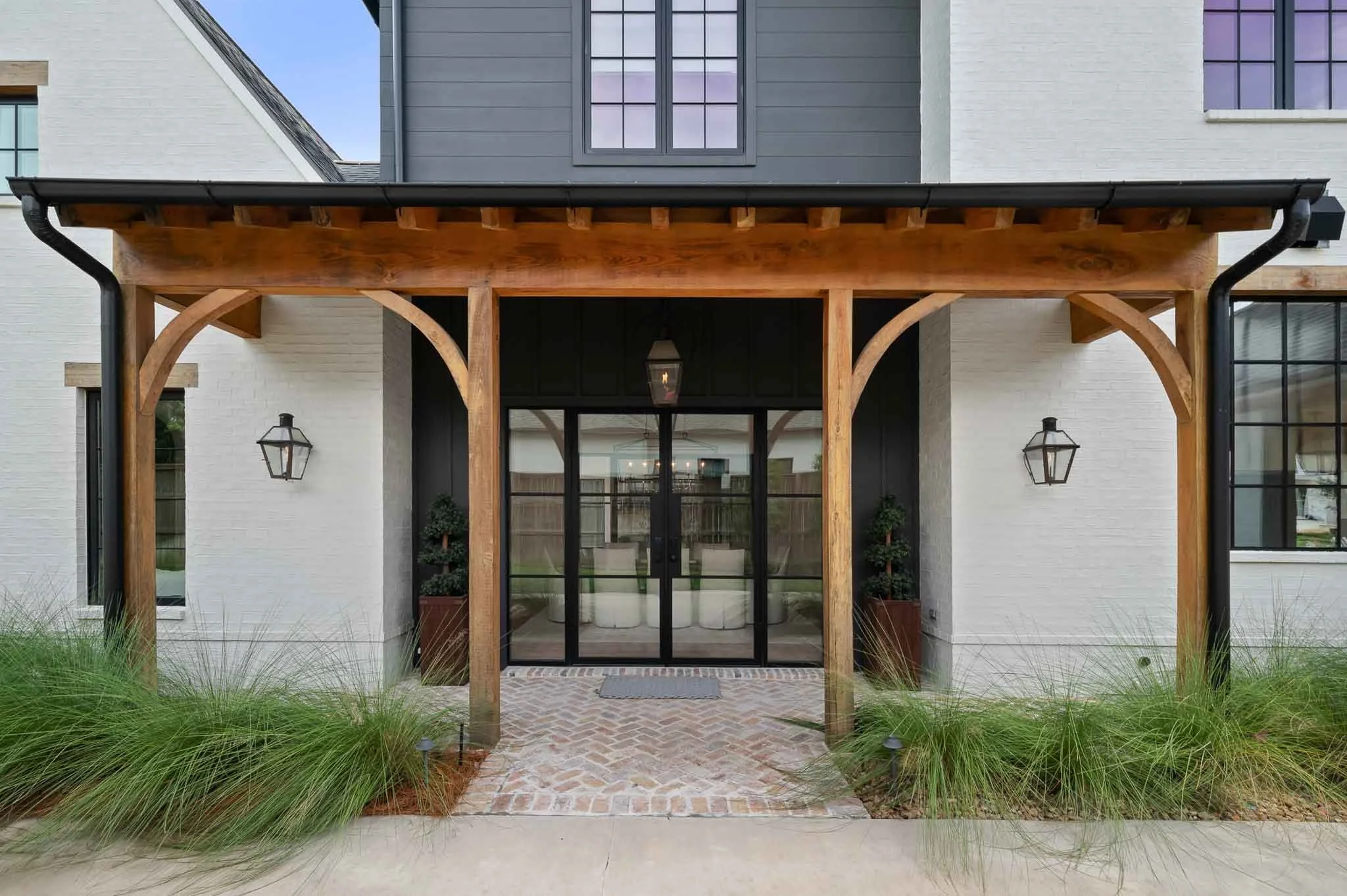 Front entrance of a house with a wooden porch, brick pathway, black glass doors, and wall-mounted lanterns, surrounded by green plants and grass.