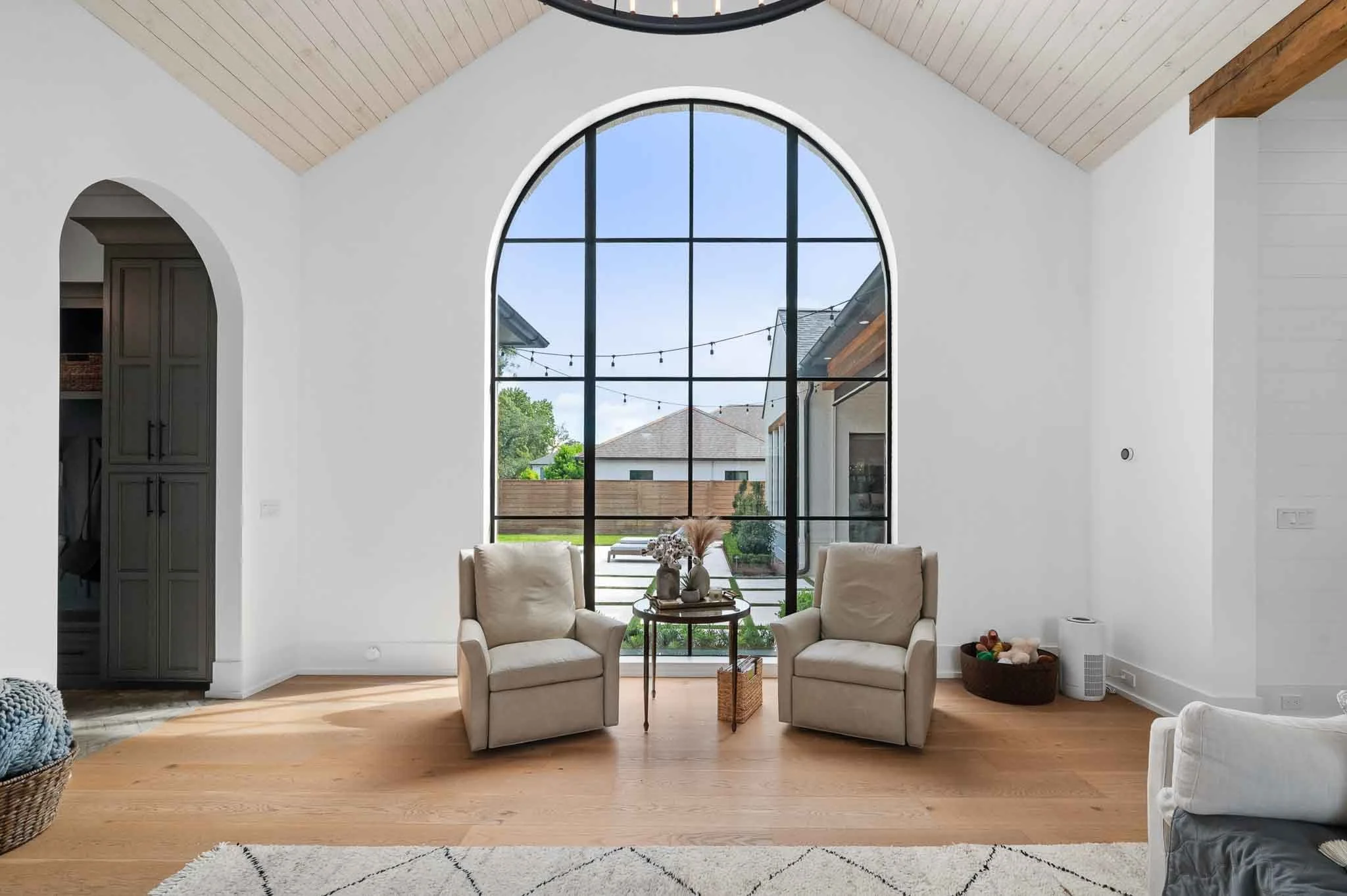 Living room with large arched window, two beige armchairs with decorative pillows, a small round table with vases and plants, wooden flooring, white walls, and a view of the backyard with string lights and fence.
