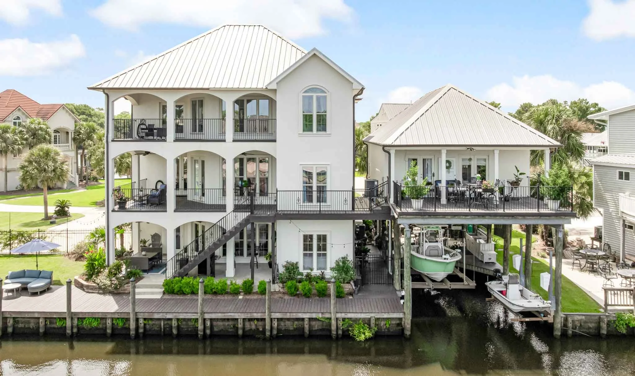 Multi-story coastal house with white exterior, multiple balconies, and a boat dock by the water, featuring outdoor seating and lush greenery.