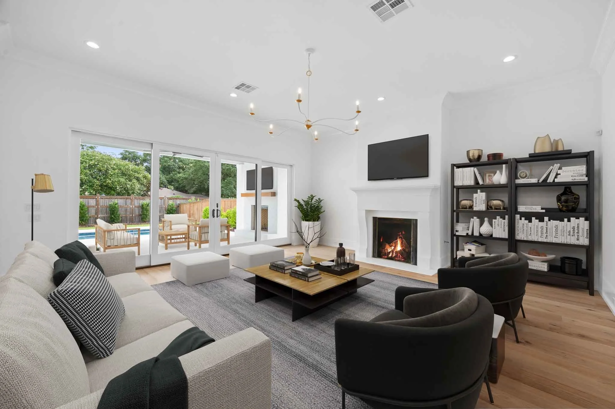 Modern living room with white walls, fireplace, large sliding glass door leading to backyard patio, black bookshelves, black armchairs, beige sofa with black and white pillows, gray rug, wooden coffee table, TV above fireplace, plants, and outdoor patio furniture in backyard.