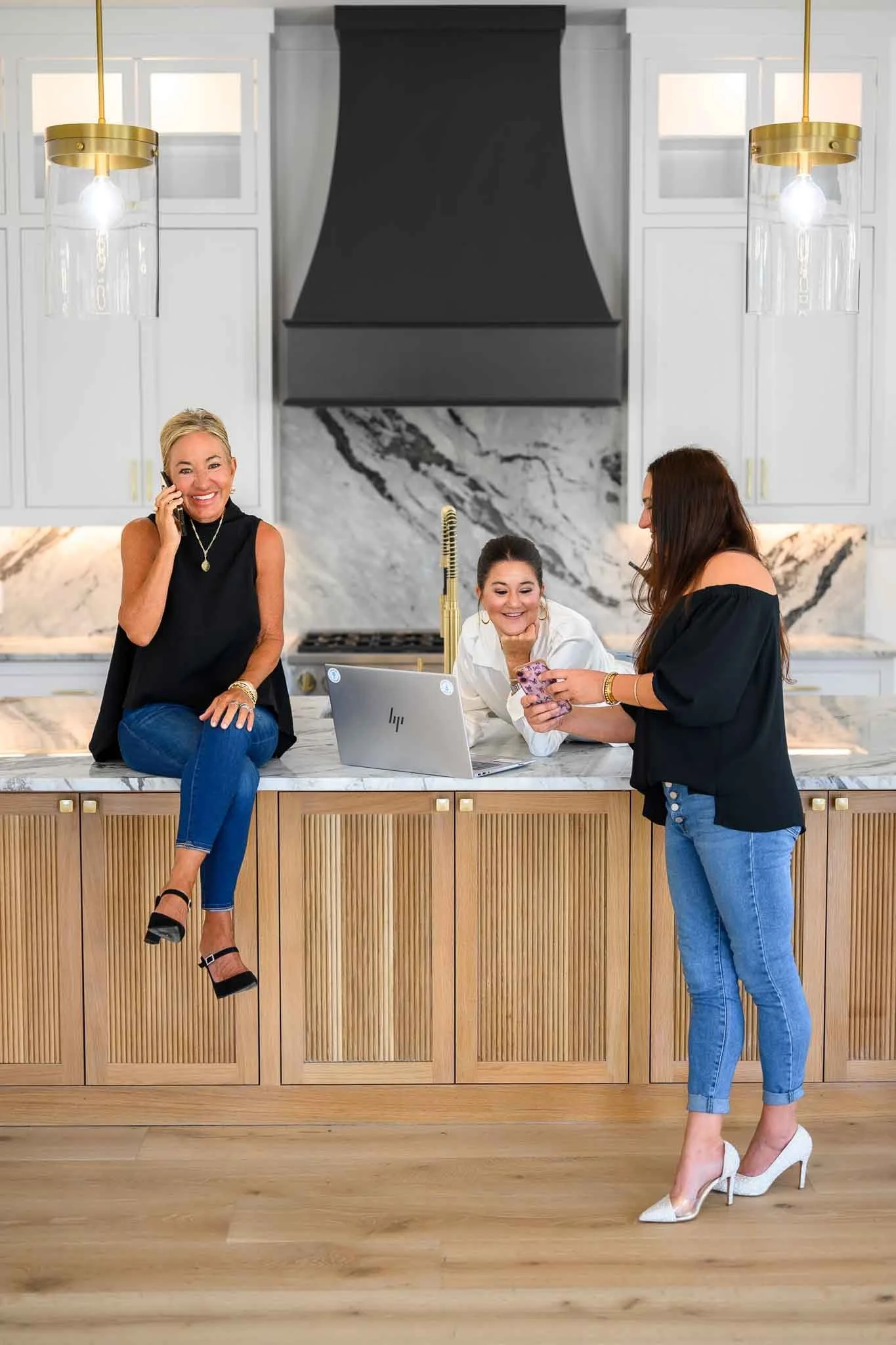 Three women in a modern kitchen, one sitting on the countertop talking on a phone, another smiling at a laptop, and the third looking at her phone, all sharing a joyful moment.