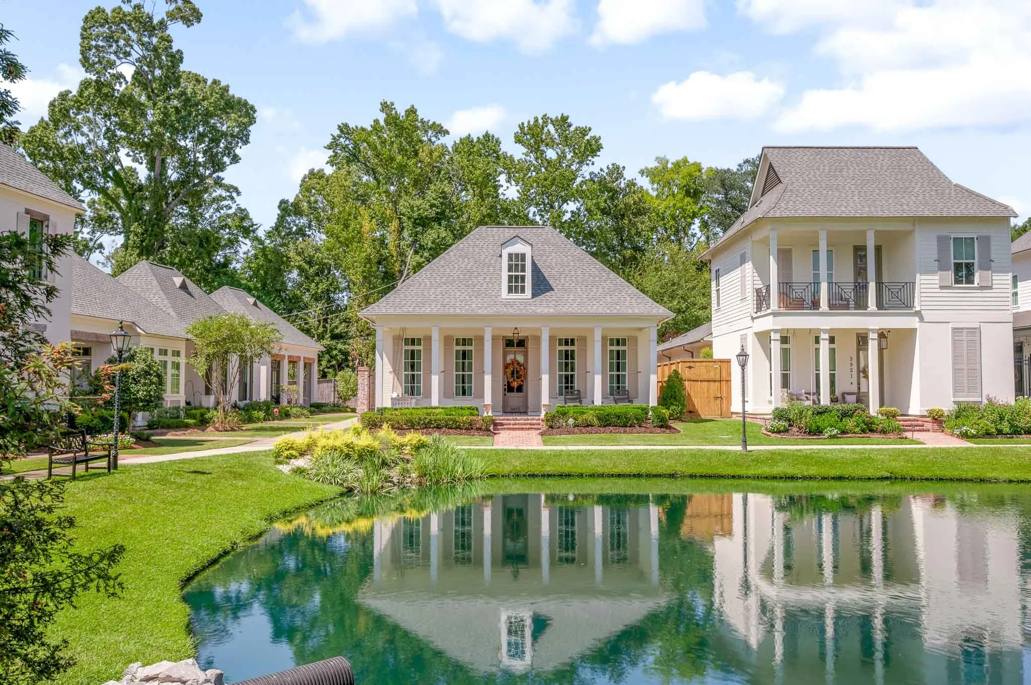 White two-story house with front porch and small balcony, surrounded by green lawn and trees, with a pond in the front reflecting the house, and neighboring houses on either side.